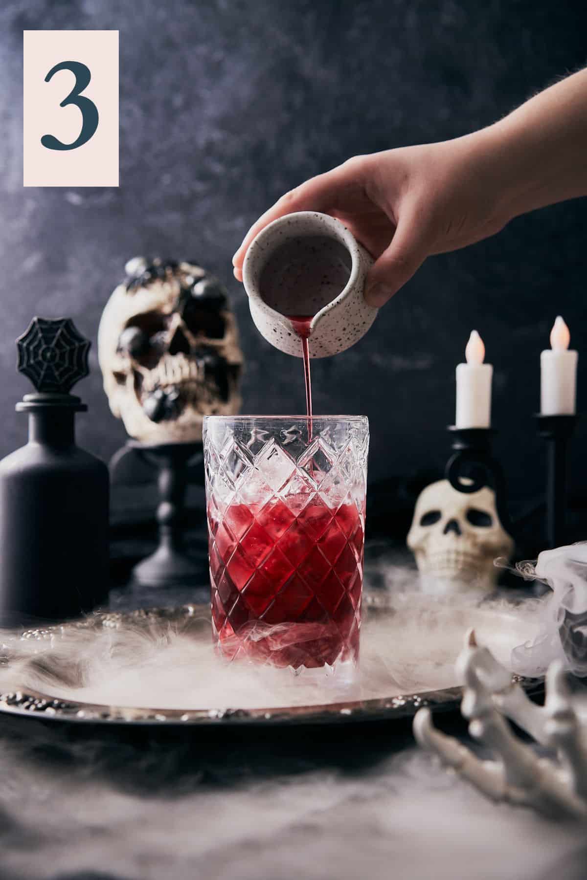 hand pouring grenadine into a glass cocktail shaker with ice in a very spooky scene with dry ice and skulls.