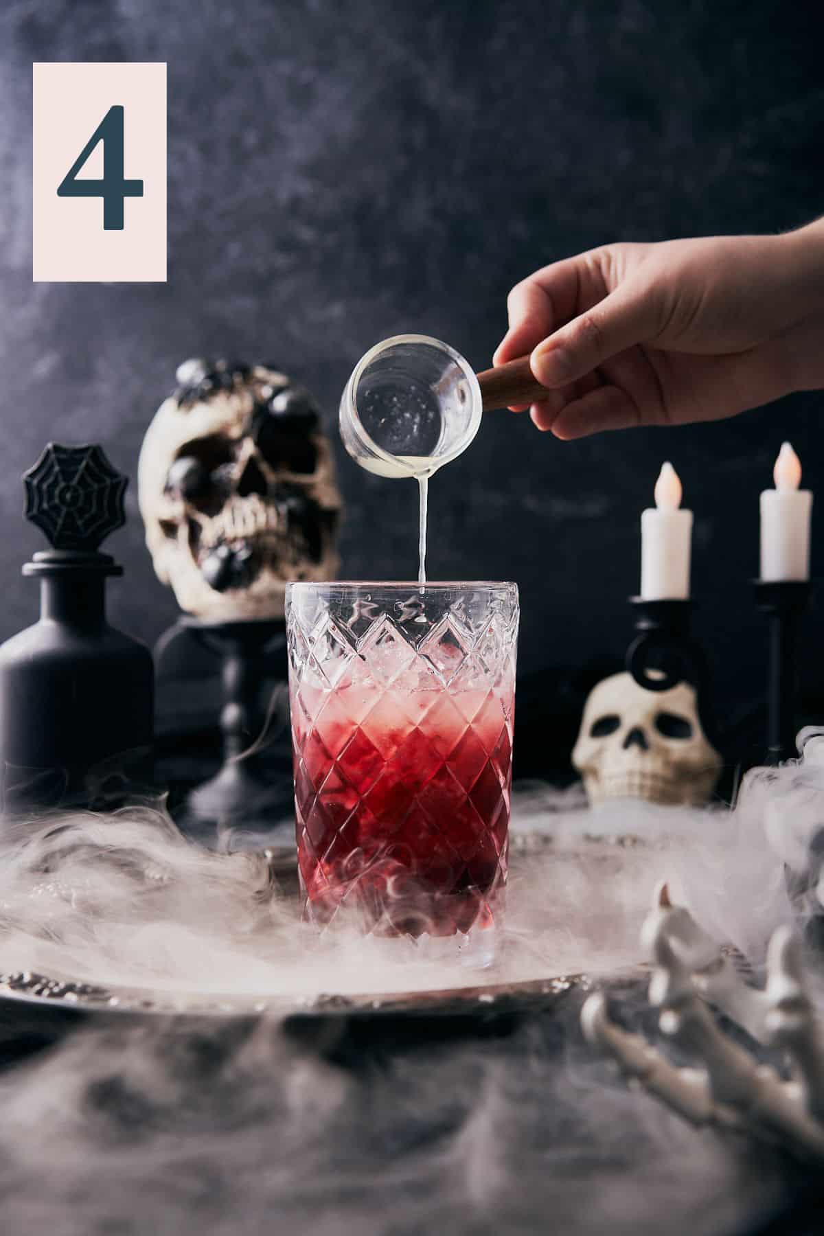 hand pouring lemon juice into a cocktail shaker with a spooky halloween scene in the background.