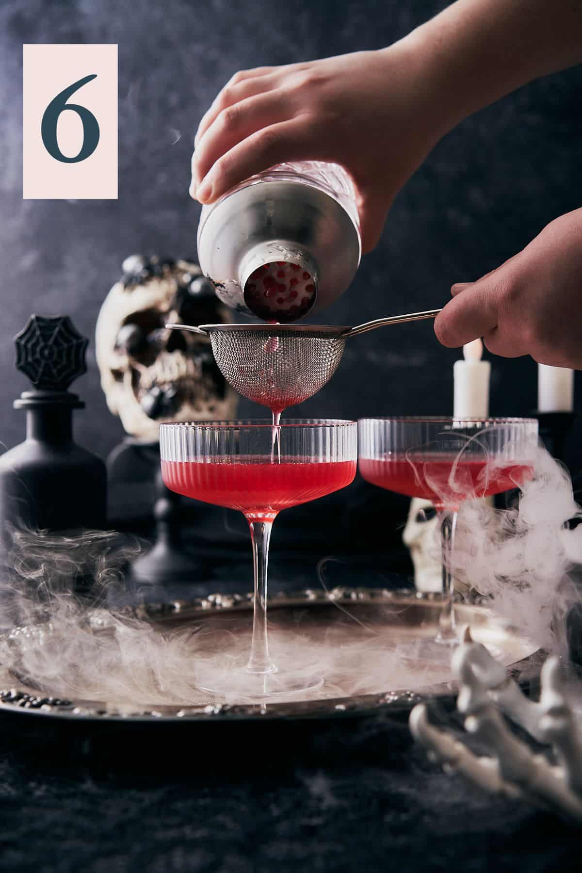 hand double straining a cocktail into a couple glass with a spooky halloween scene surrounding it and dry ice.