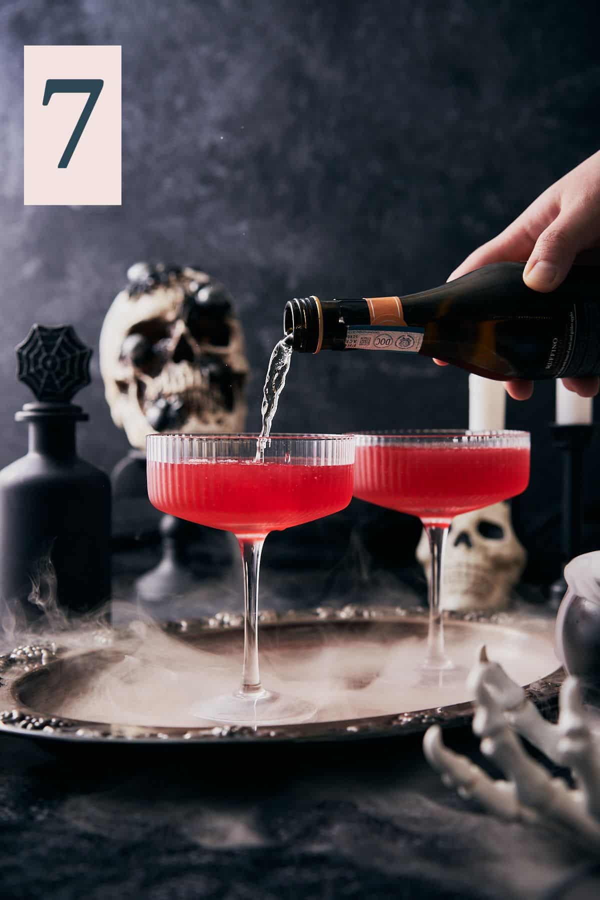 hand pouring sparkling wine into coupe glasses with red liquid inside, on a spooky dark halloween scene with skulls and dry ice.