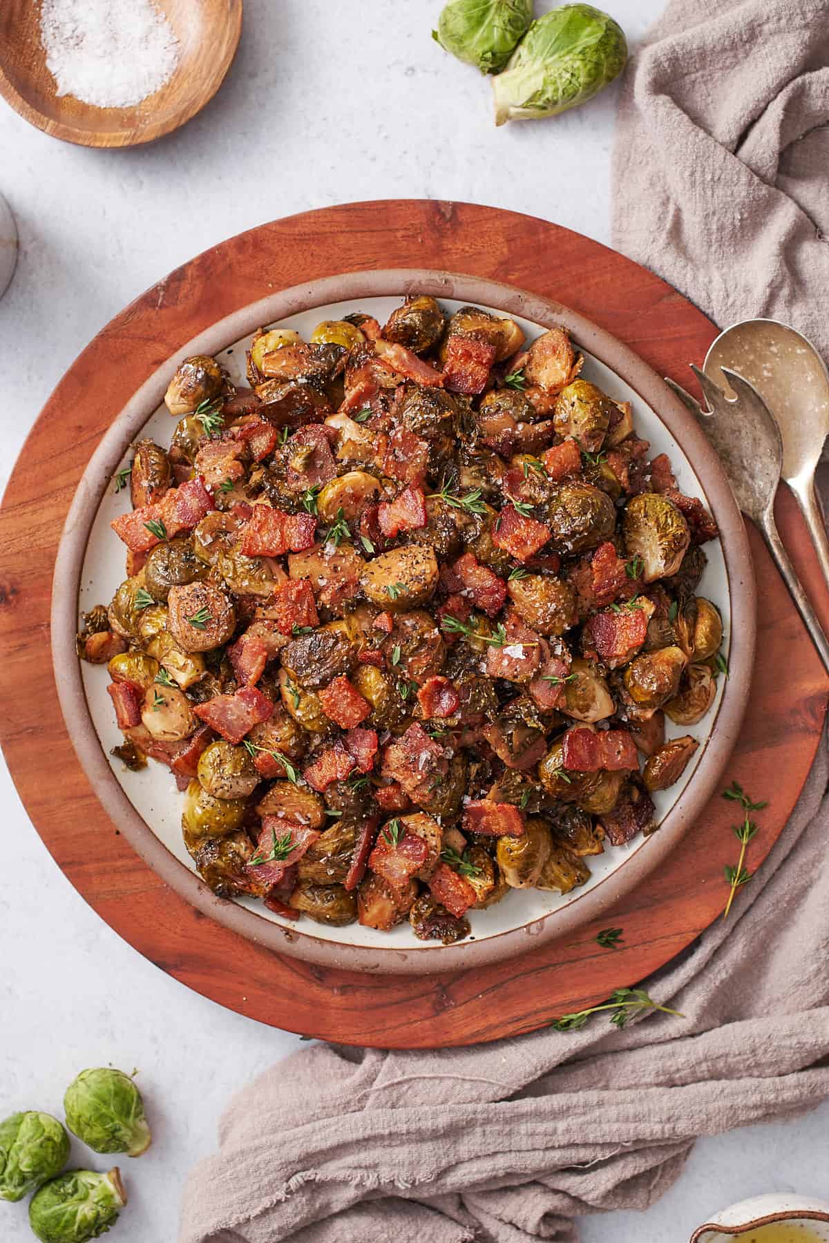 cirspy bacon balsamic brussels sprouts on a plate on a wooden serving board, serving utensils, and topped with thyme. 
