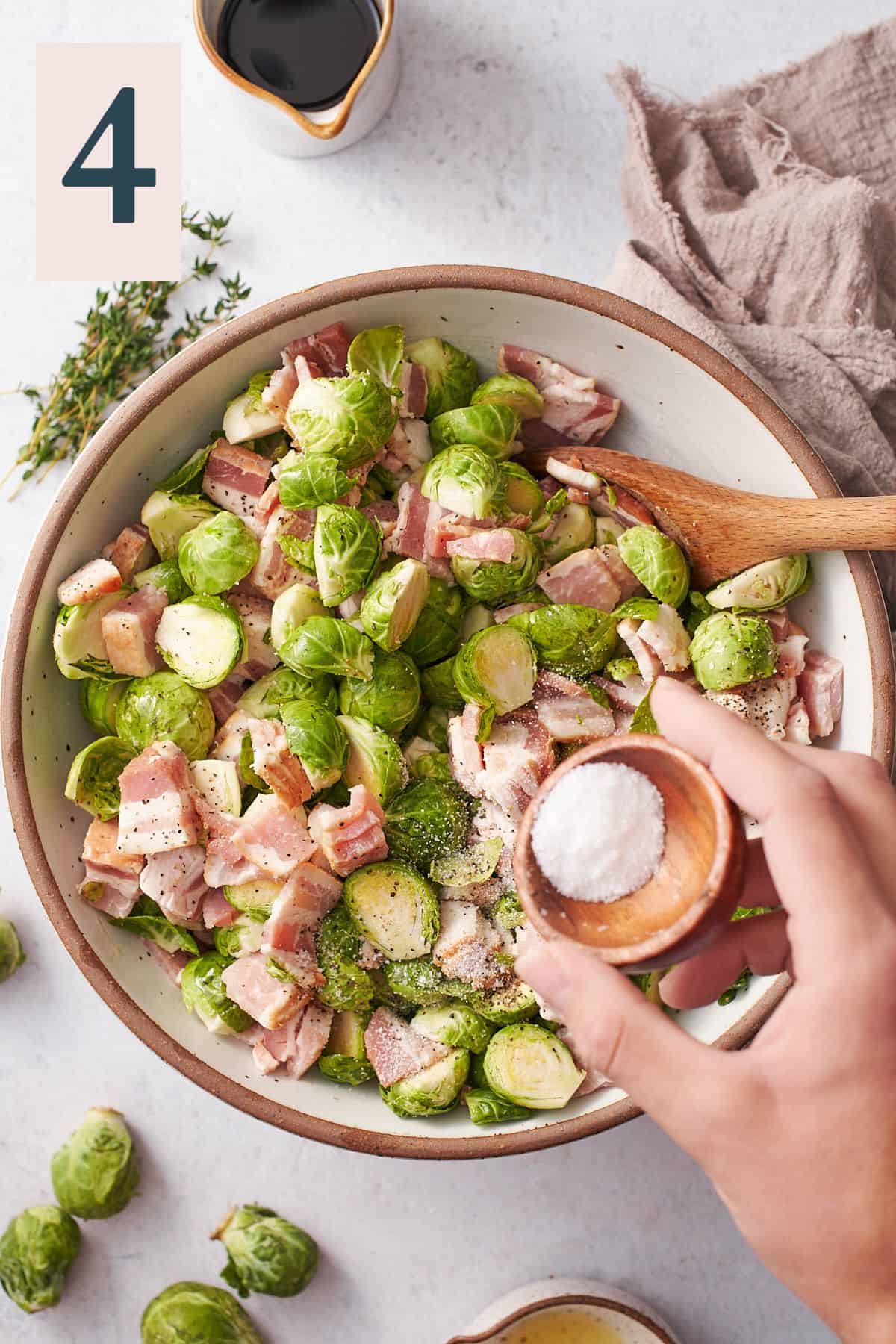 hand pouring kosher salt into a large bowl of bacon and brussels sprouts. 