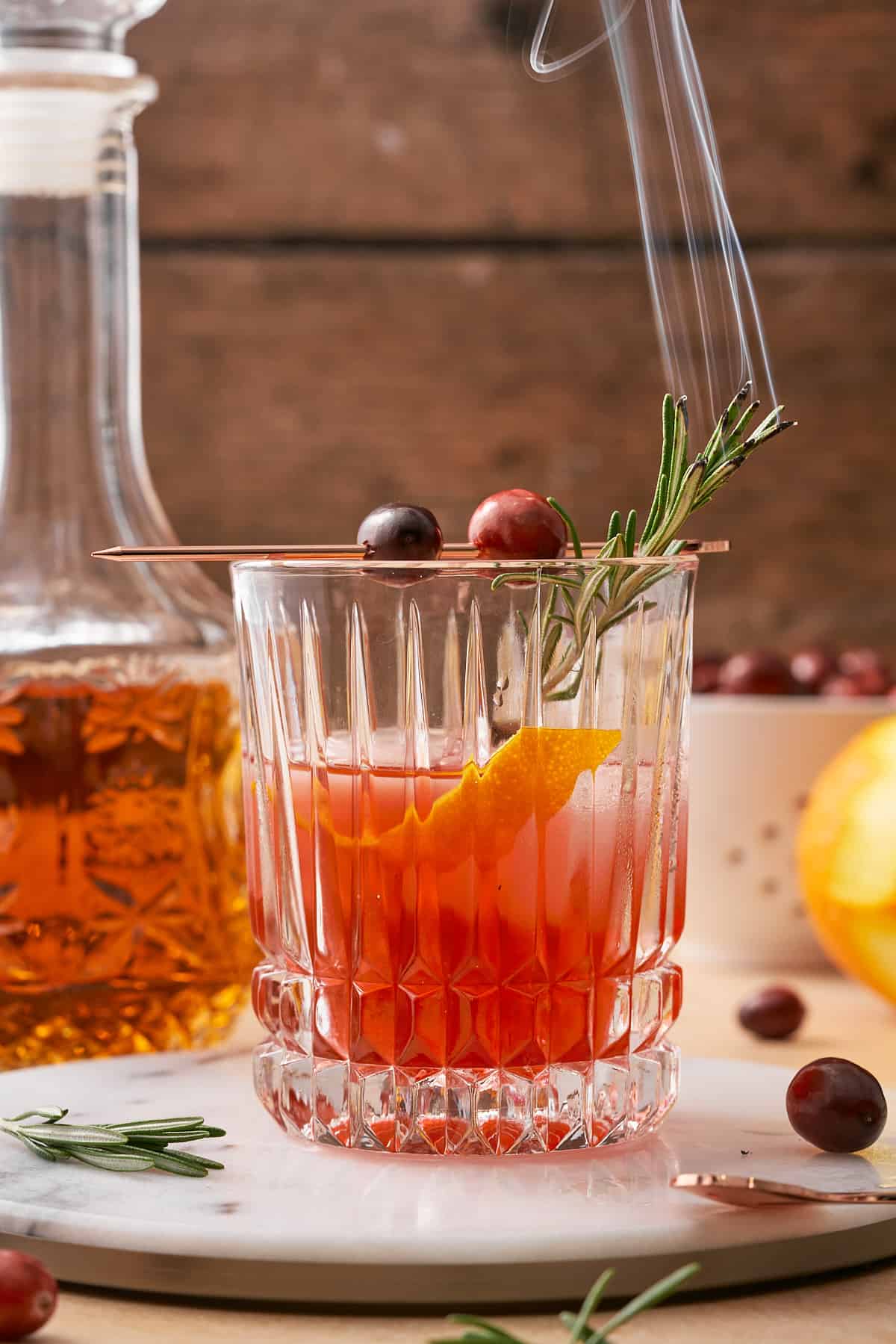 cranberry old fashioned garnished with an orange twist, smoking rosemary, and fresh cranberries. 
