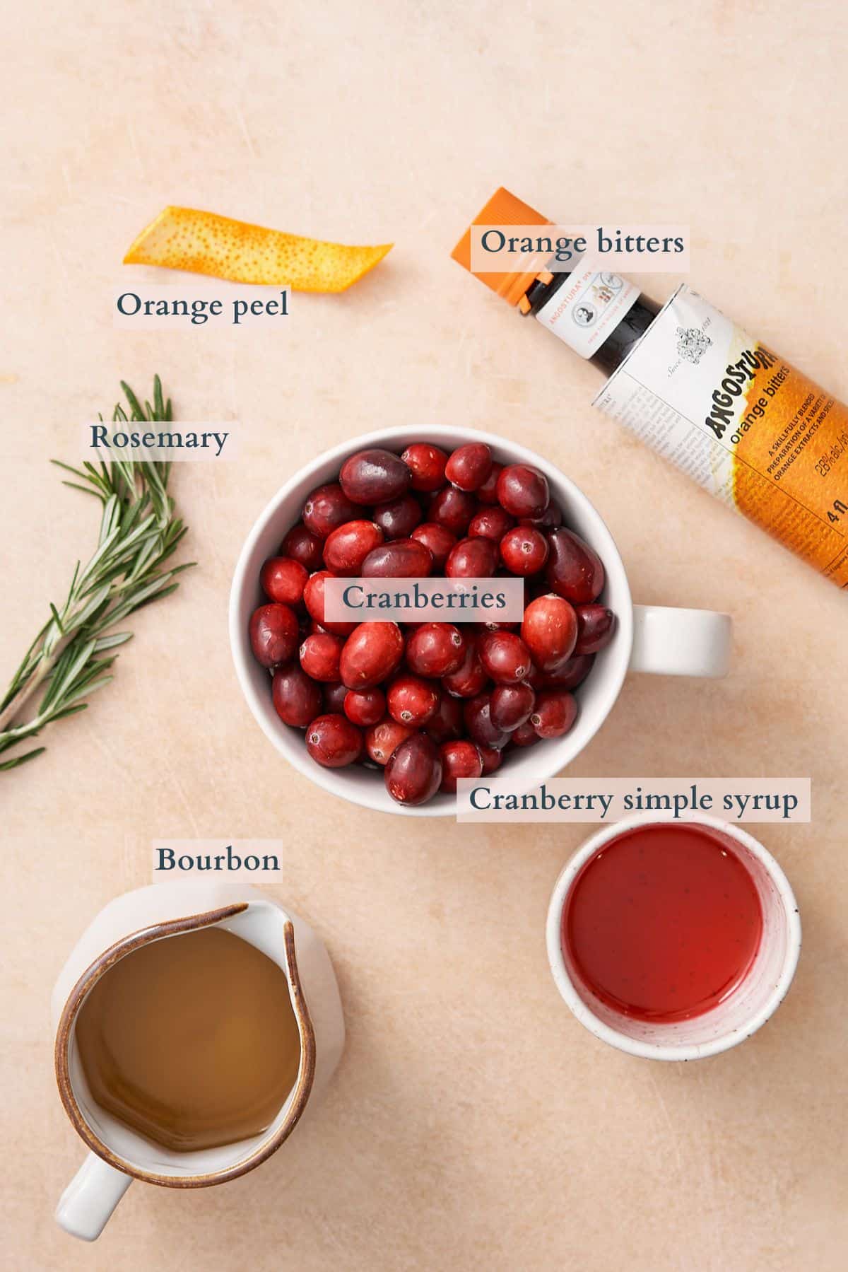 ingredients to make a cranberry old fashioned, with ingredients in separate ceramic bowls and pitchers, with text denote each ingredient. 