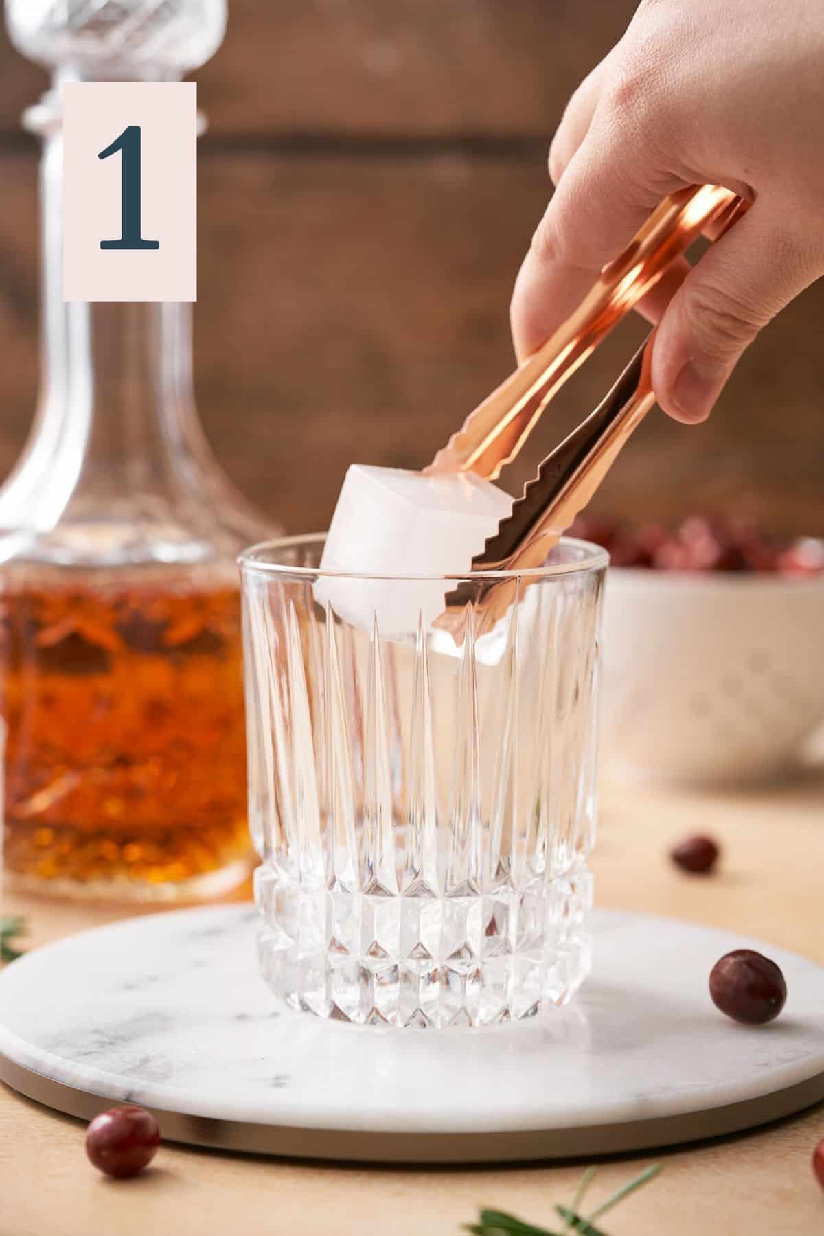 Hand using an ice tongs to place a large square ice cube into a rocks glass, with bourbon and cranberries in the background.