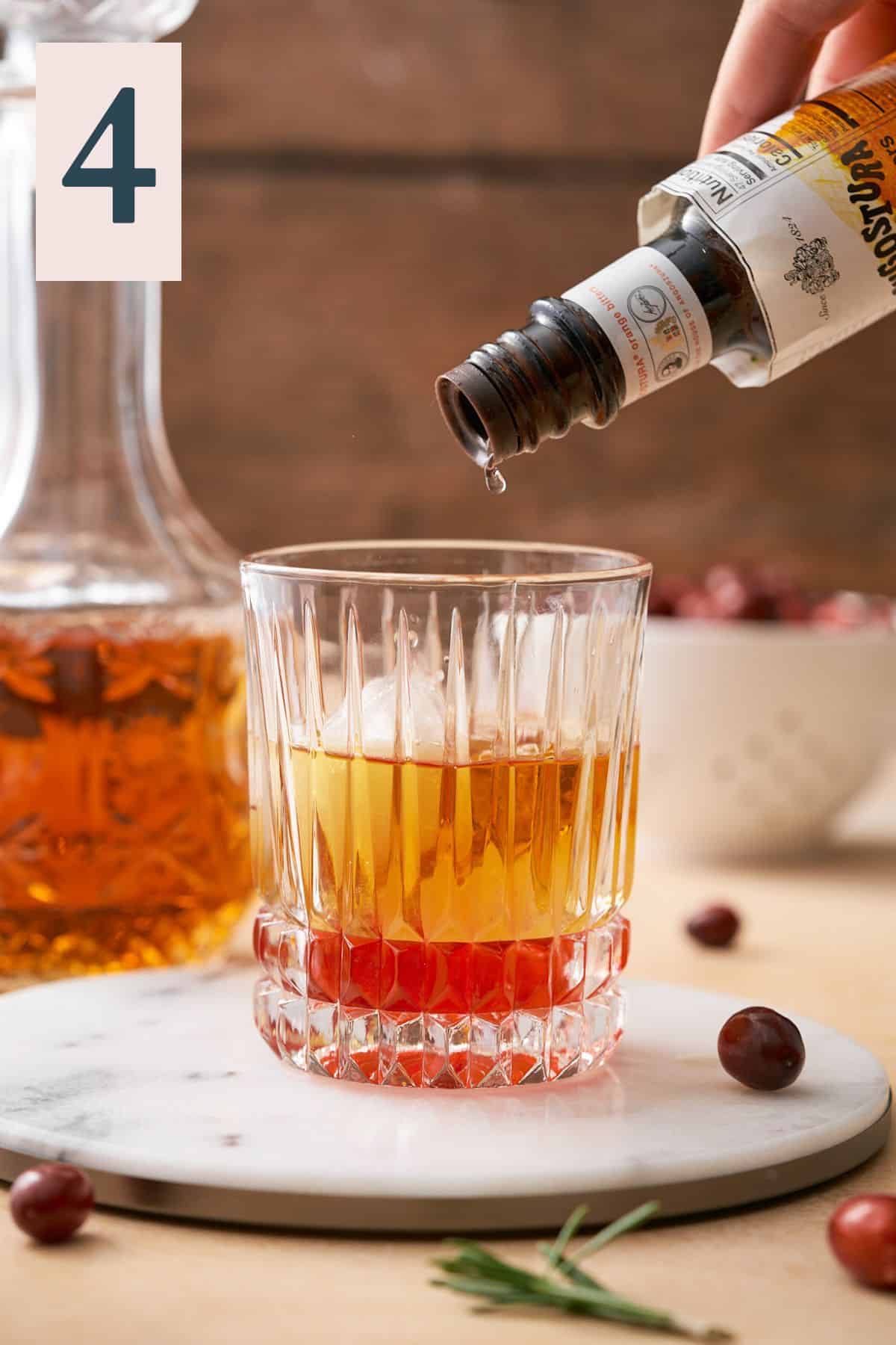 pouring angostura orange bitters into a glass with ice, bourbon, and cranberry simple syrup. 