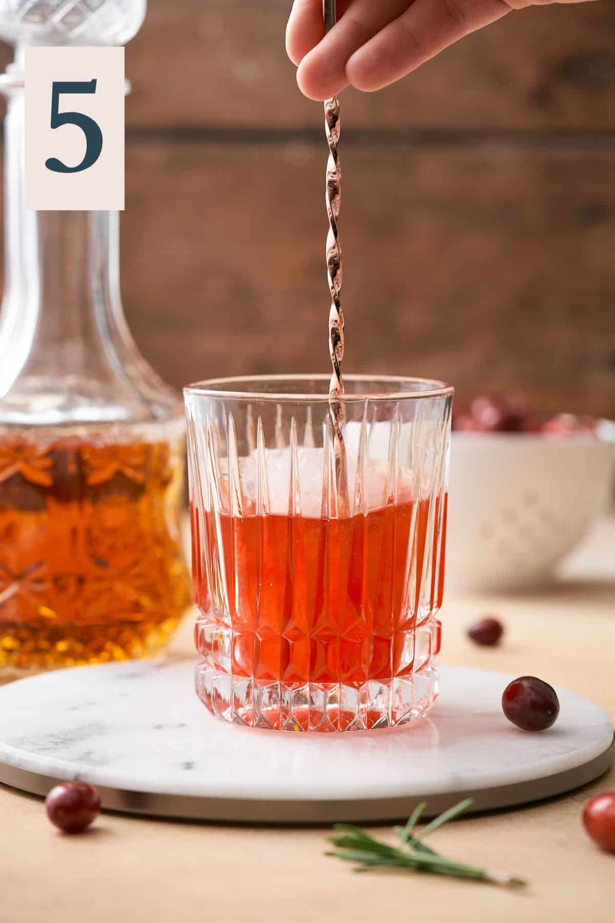 stirring a cranberry old fashioned in a rocks glass with a rose gold bar spoon. 