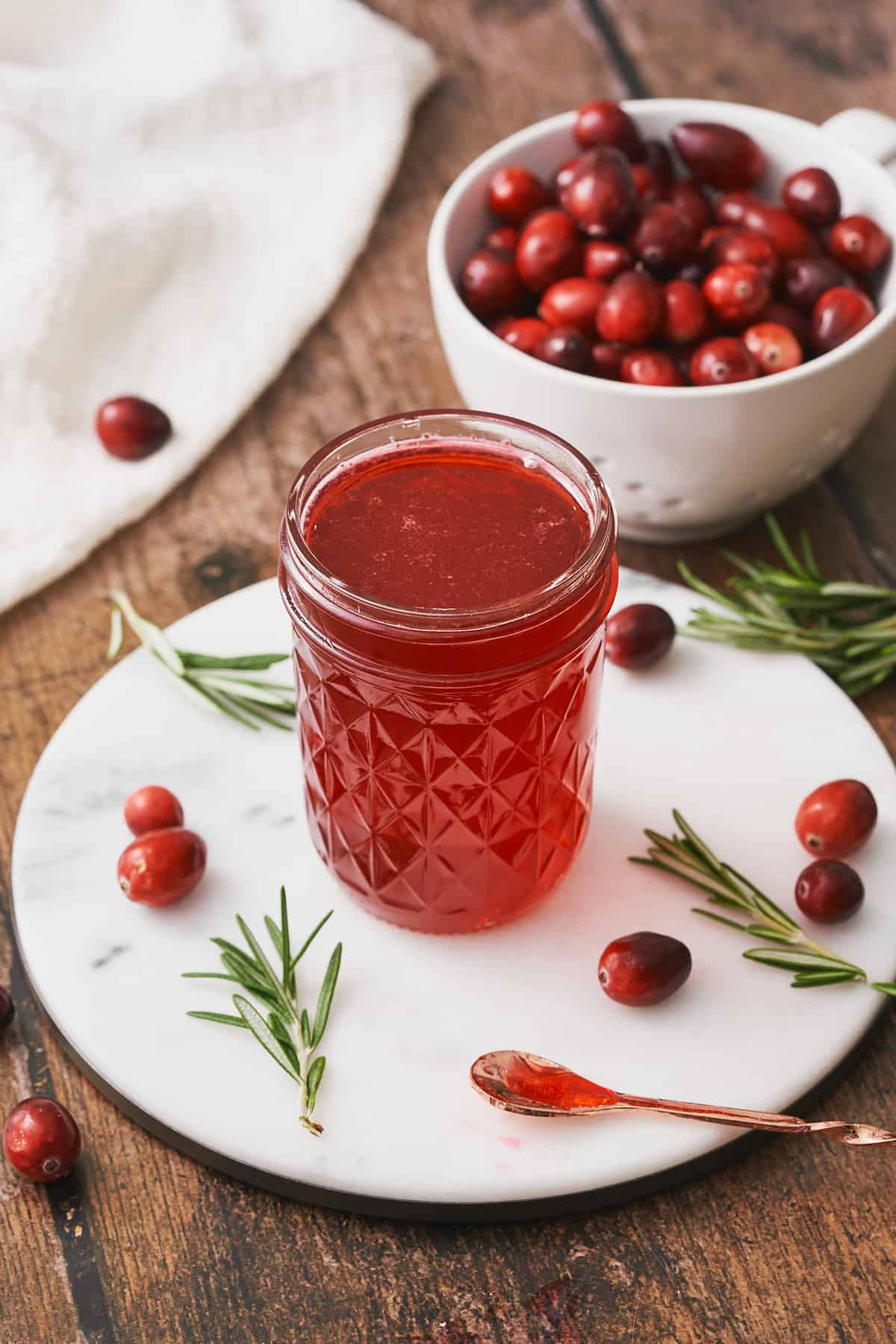 cranberry simple syrup in a glass jar on top of a round white marble heat plate, surrounded by cranberries, and fresh rosemary.  