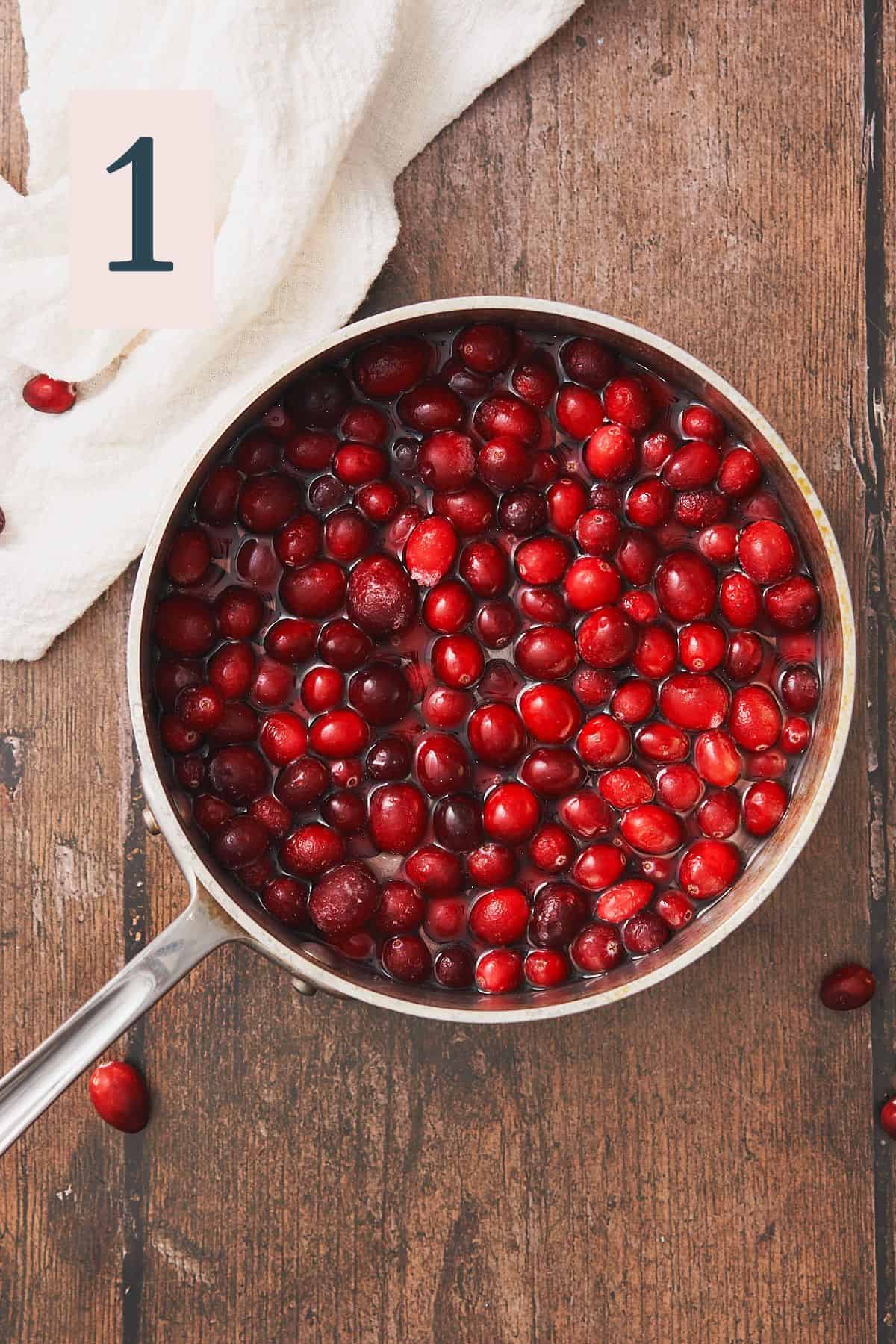 cranberries in a saucepan with water. on a dark wood board, with fresh cranberries surrounding the pan.