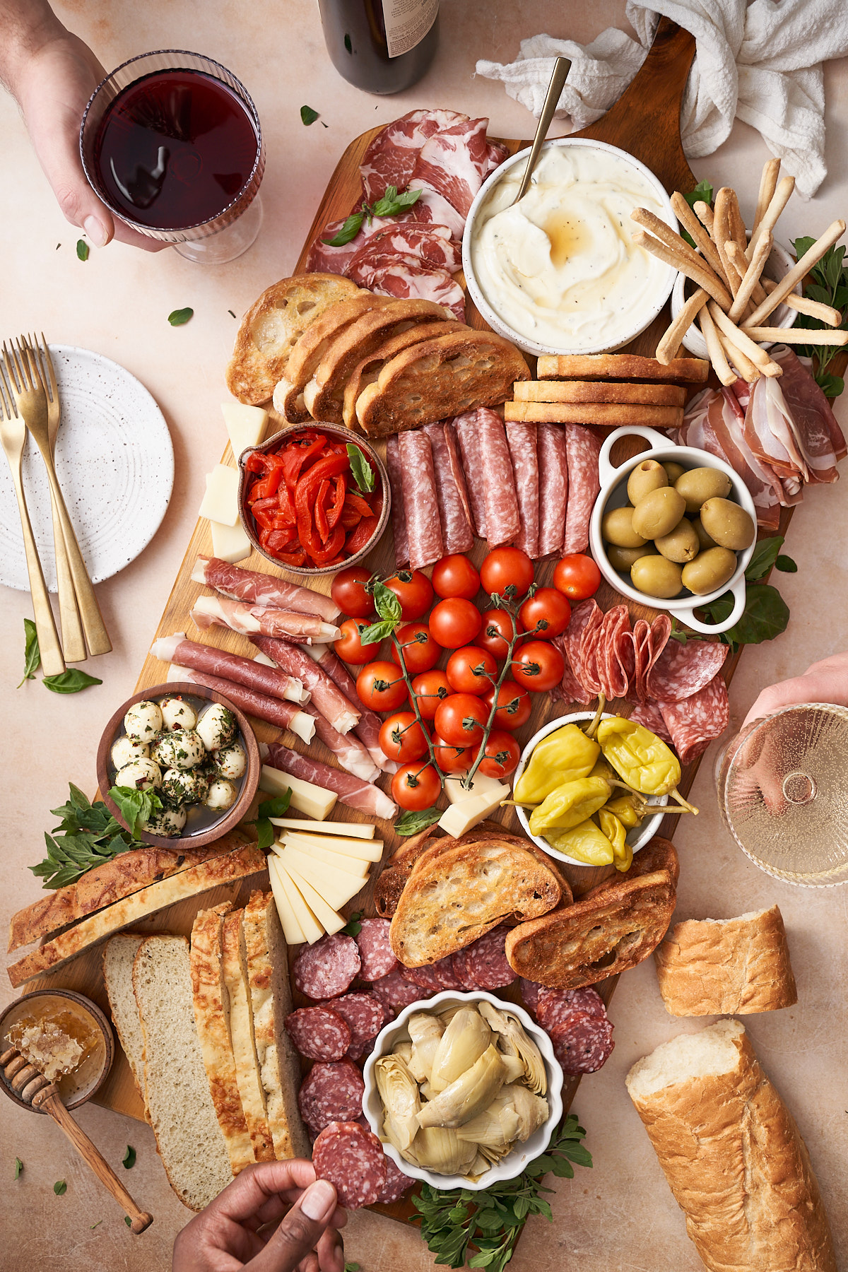 large antipasto platter with bread, cherry tomatoes, marinated vegetables, cheese, and hands reaching for wine and snacks on the board.