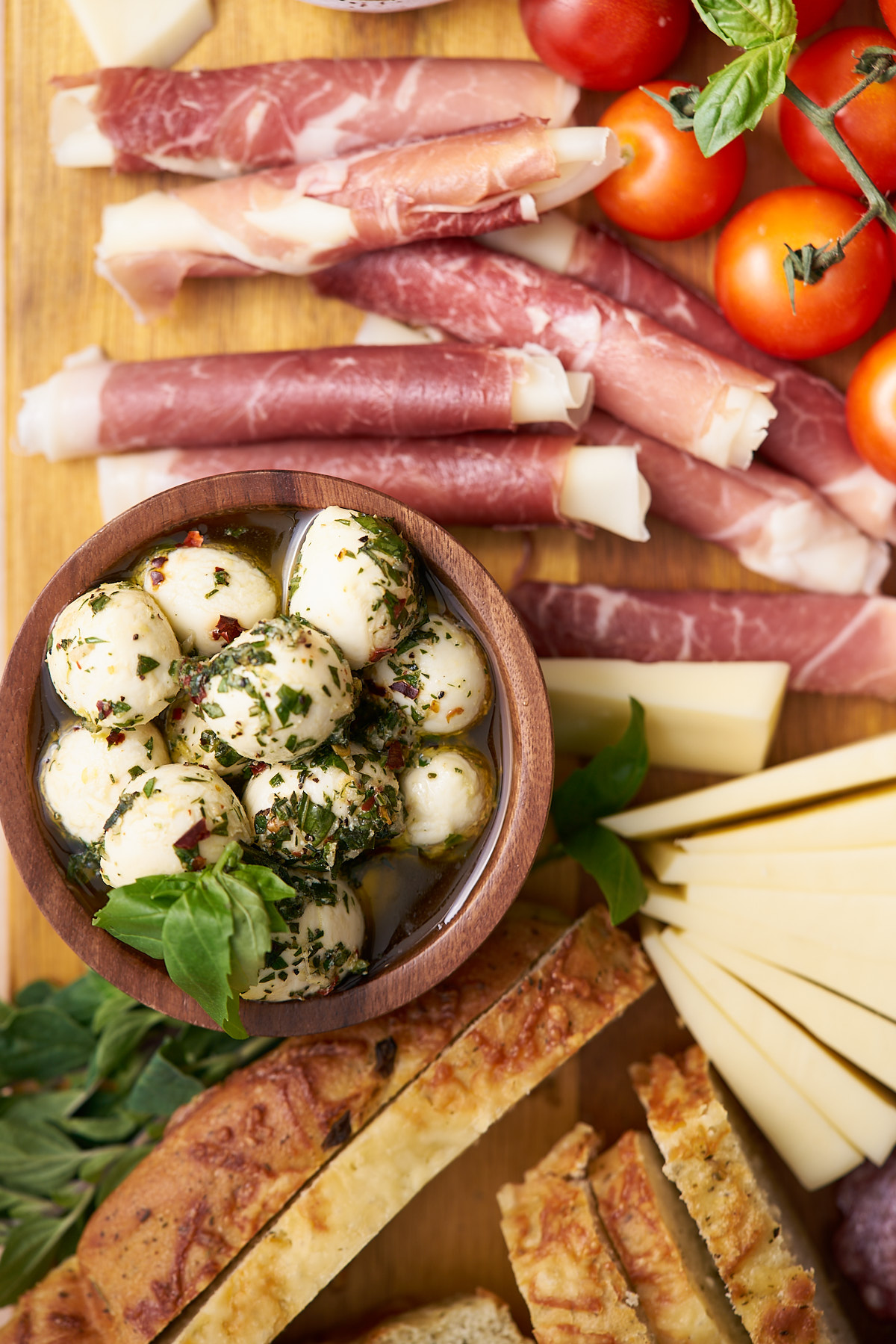 marinated mozzarella balls on an Italian antipasto platter with prosciutto, focaccia, cherry tomatoes, fresh herbs, and provolone cheese.