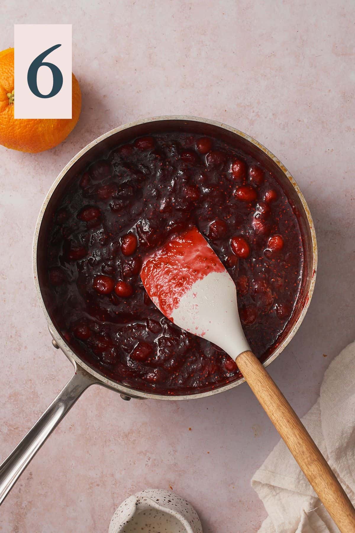 cranberry sauce thickening in a saucepan with a rubber spatula, and an orange nearby.