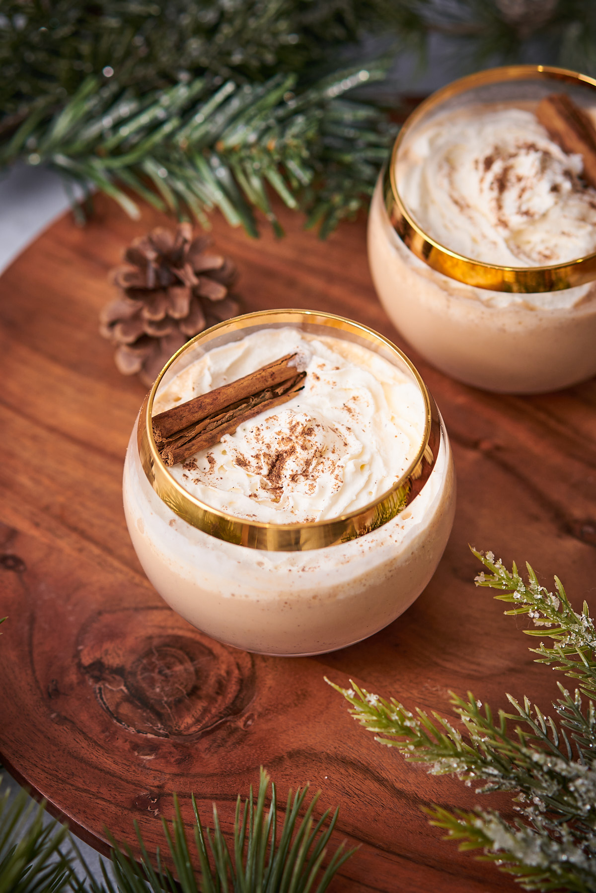 keto eggnog in round glasses, with a gold rim, then topped with whipped cream, nutmeg, and a cinnamon stick, with balsam and a pinecone in the background. 