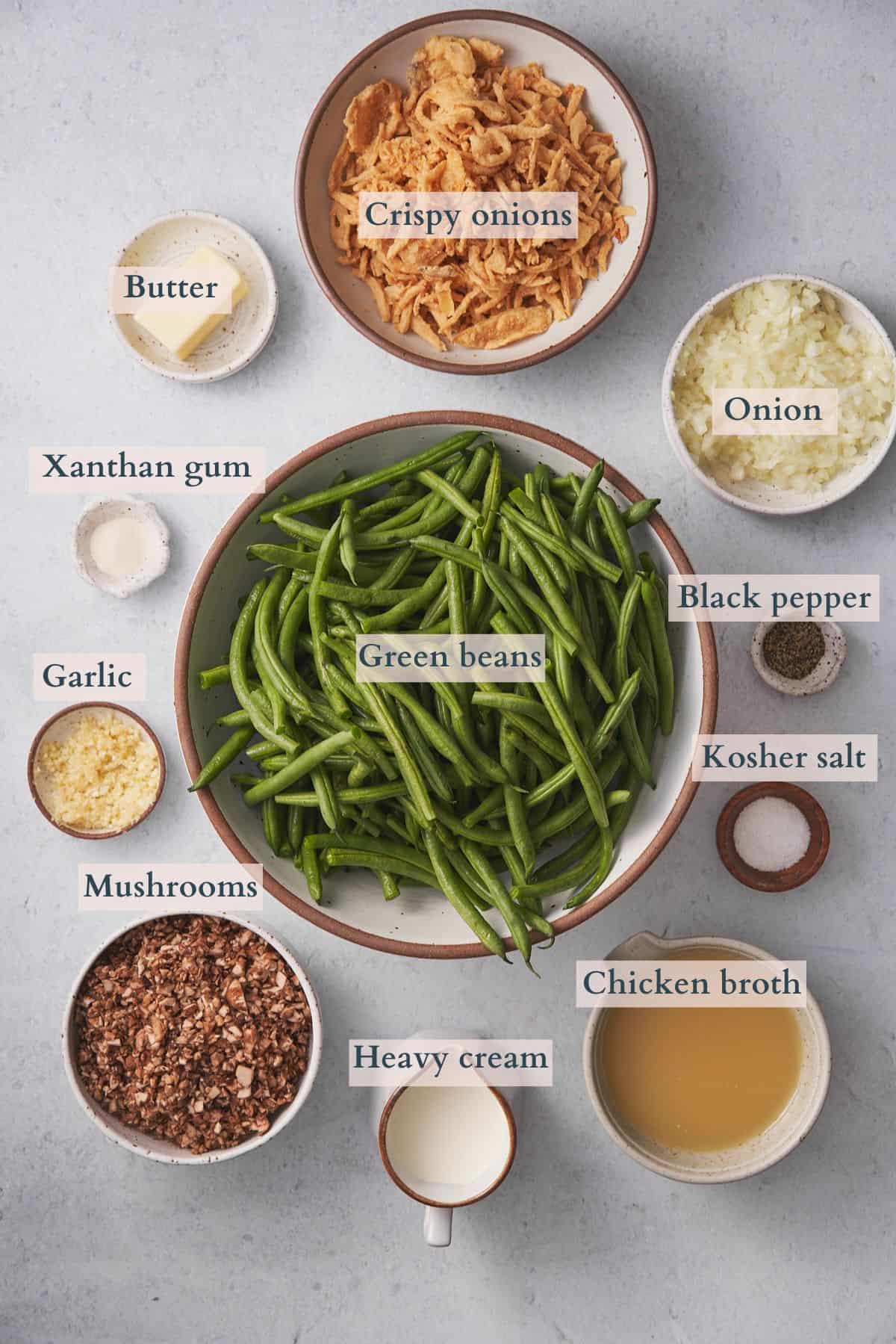 ingredients to make keto green bean casserole laid out in ceramic bowls with text overlaying to denote each ingredient. 