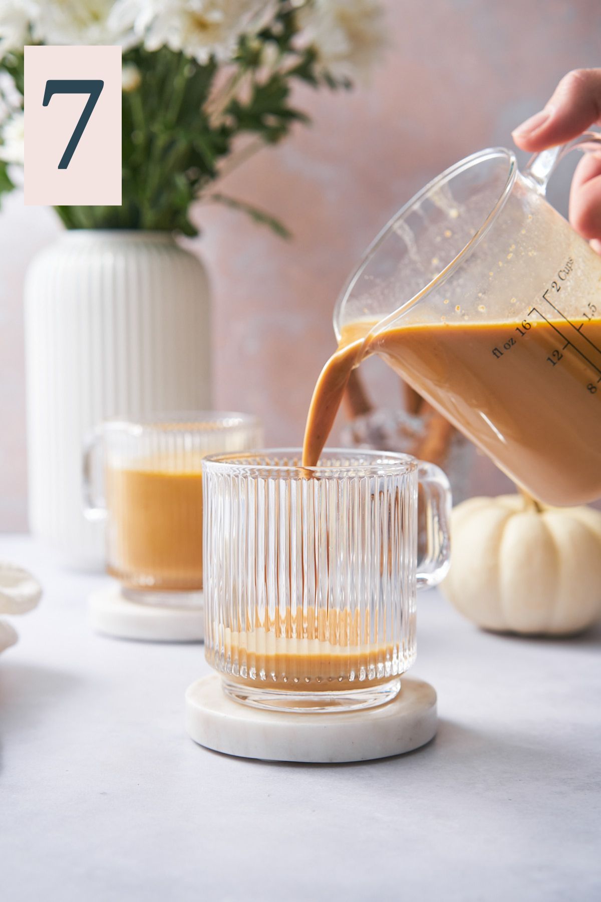hand pouring pumpkin spice latte mixture into glass mugs with pumpkins in the background, along with white mums and cinnamon sticks. 