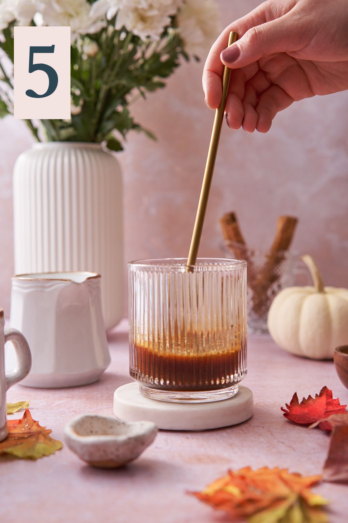 hand stirring coffee in a glass mug surrounded by maple leaves, pumpkins, and white mums in the background. 