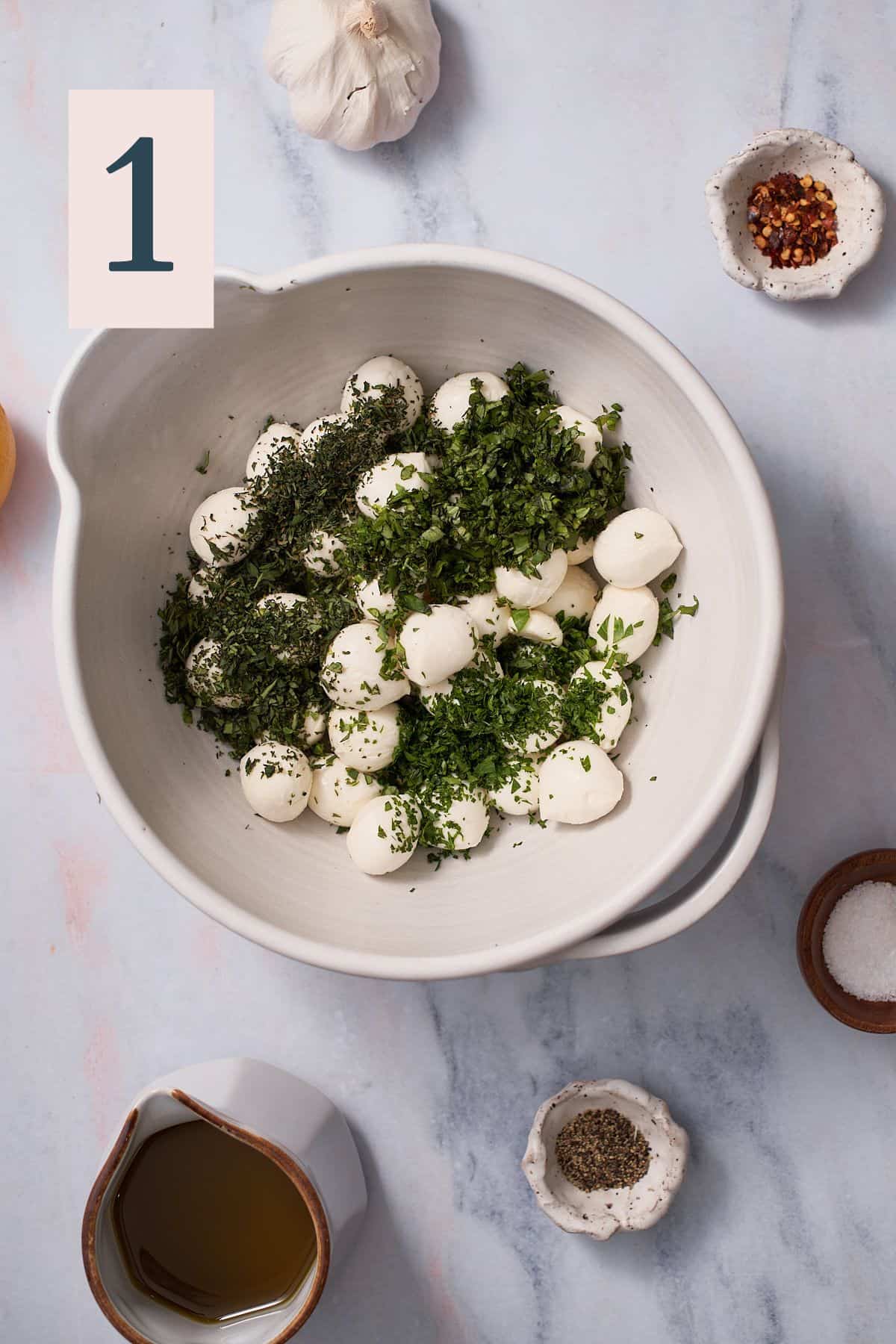 mozzarella balls in a large bowl with fresh minced herbs, surrounded by red pepper flakes, kosher salt, black pepper, olive oil, and lemon.