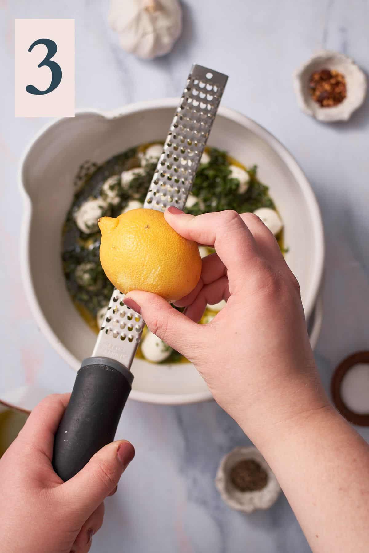 hand zesting lemon zest into a bowl of mozzarella balls with herbs and olive oil. 