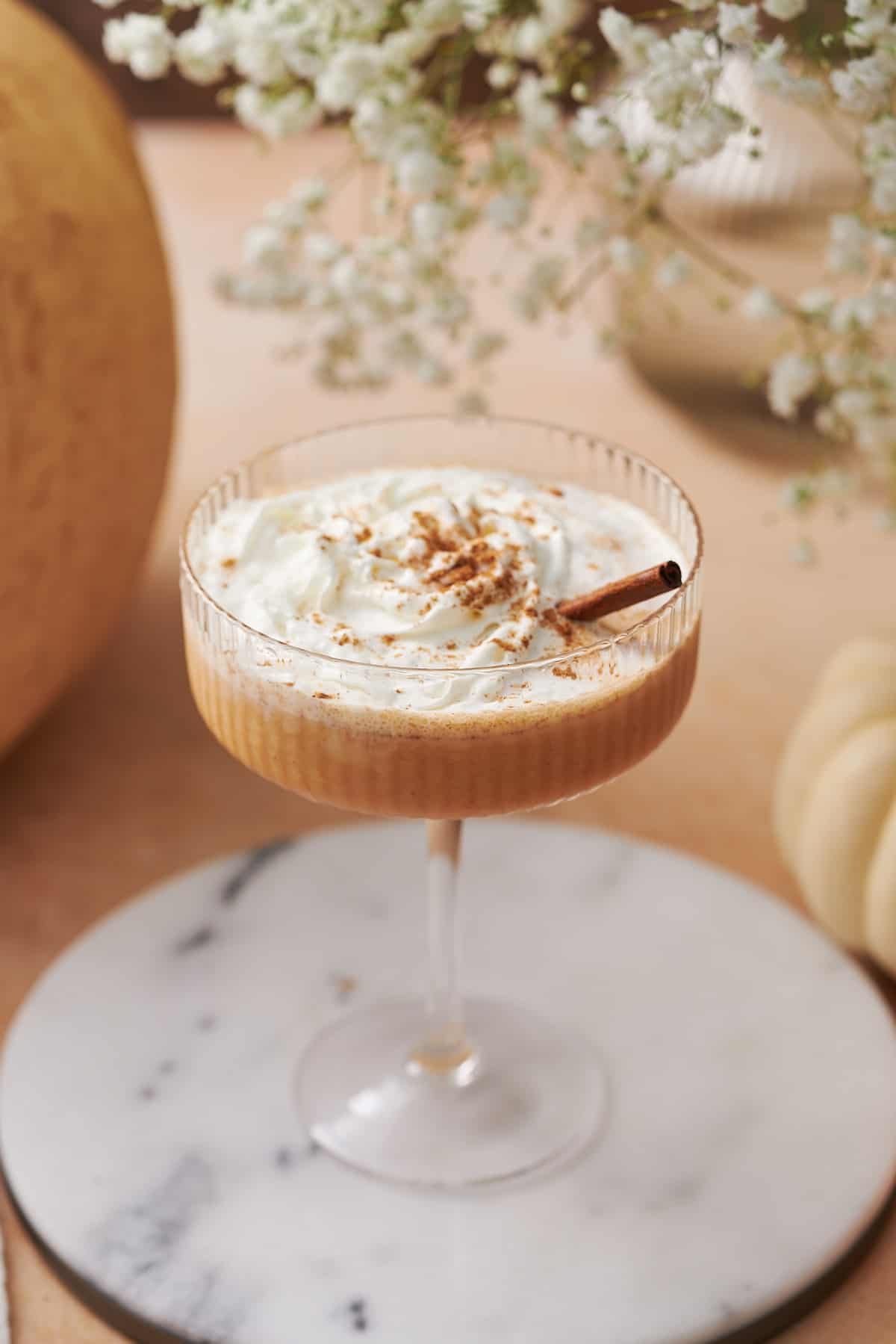 close up shot of a pumpkin martini topped with whipped cream, a cinnamon stick, and pumpkin pie spice, with little white flowers, and different gourds in the image. 