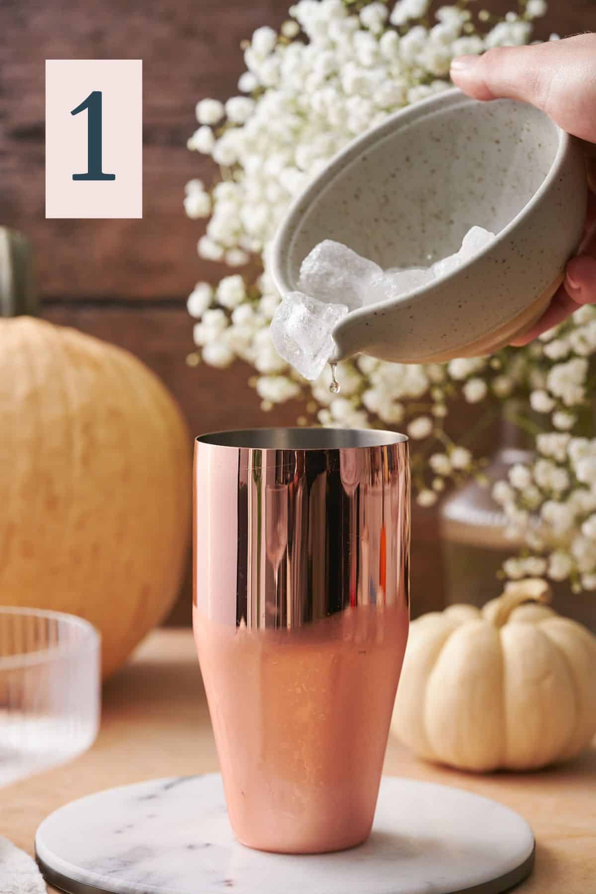 hand pouring ice into a cocktail shaker, with white flowers and various gourds in the background. 