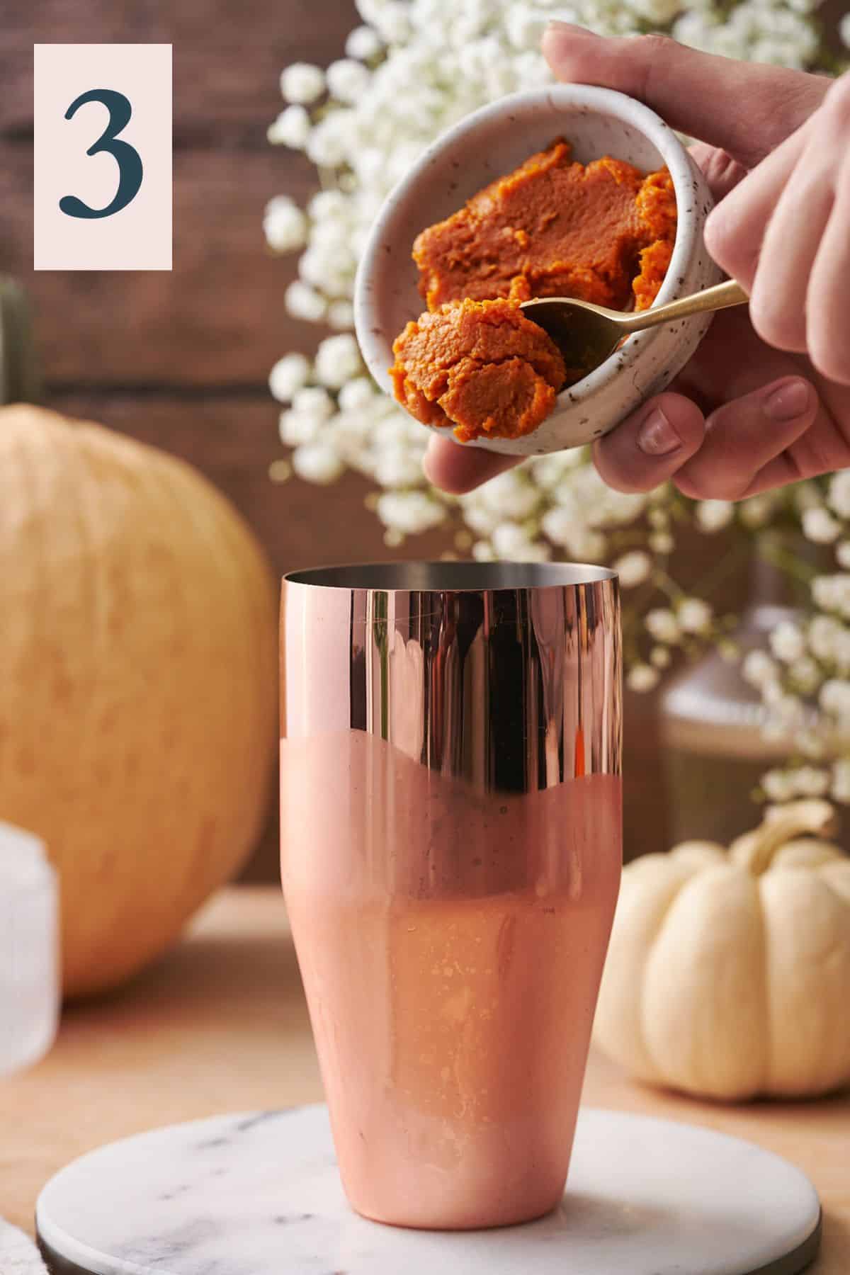 hand pouring pumpkin puree into a cocktail shaker, with white flowers and various gourds in the background. 