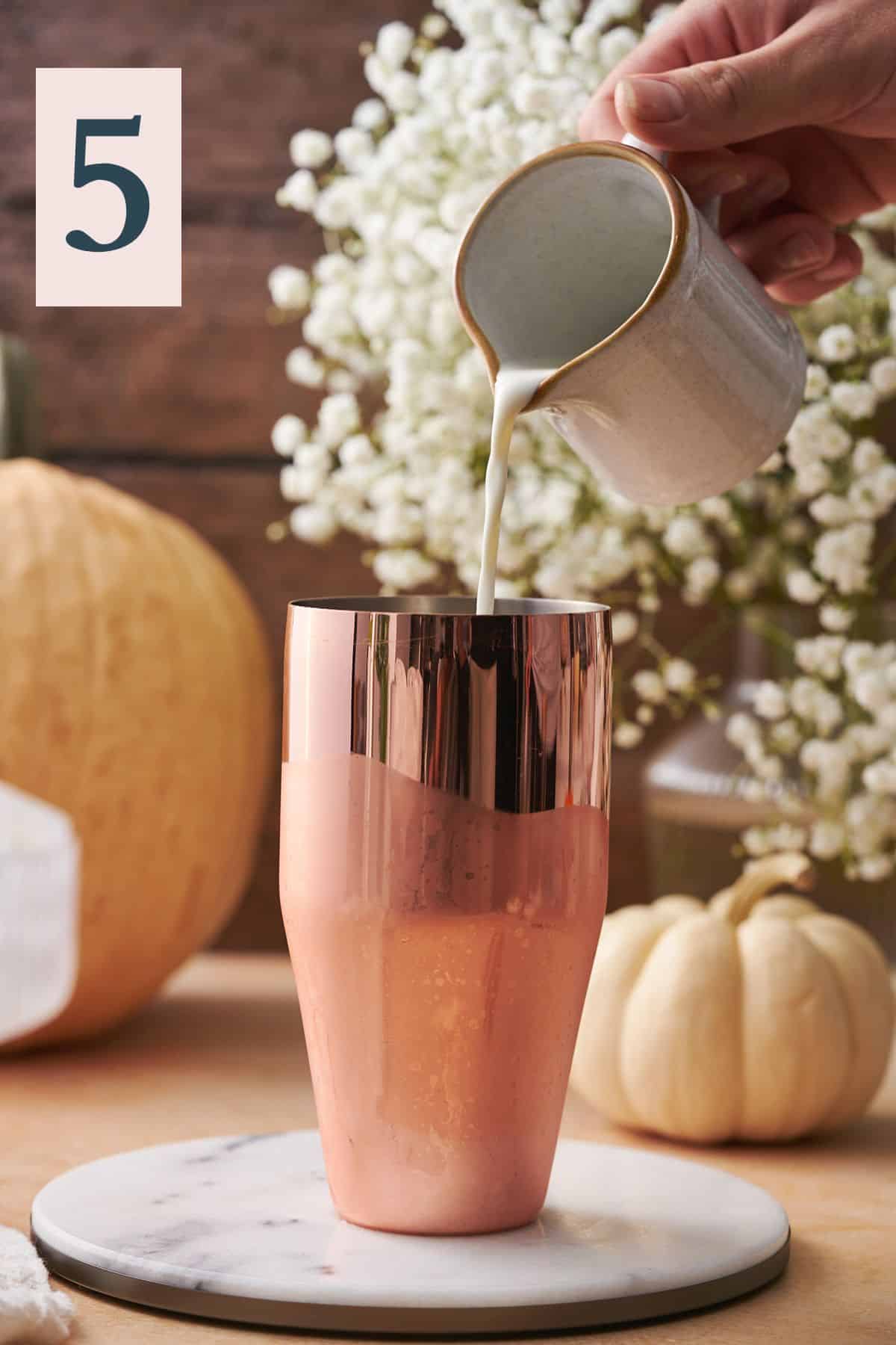 hand pouring half and half into a cocktail shaker, with white flowers and various gourds in the background. 