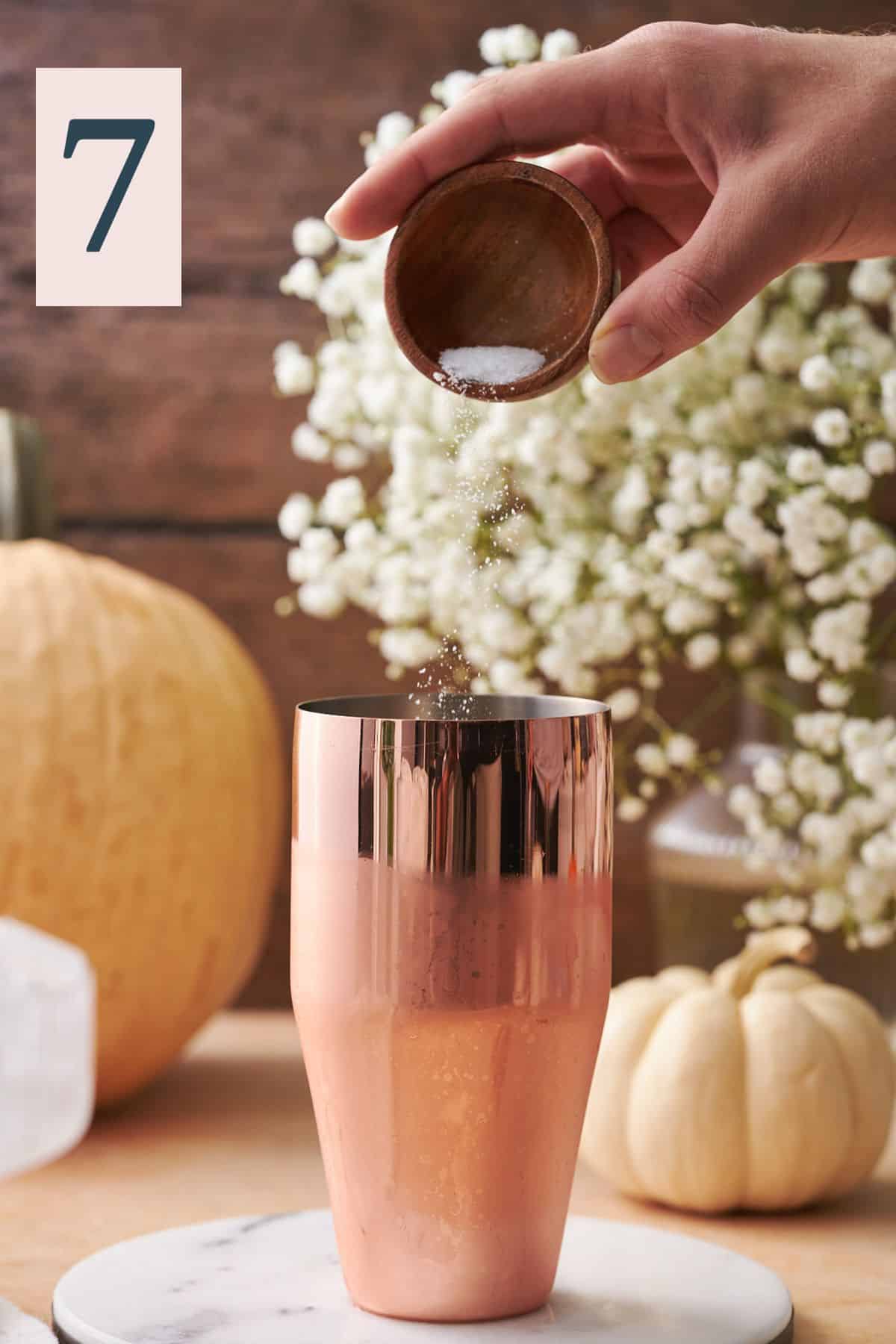 hand adding salt into a cocktail shaker, with white flowers and various gourds in the background. 