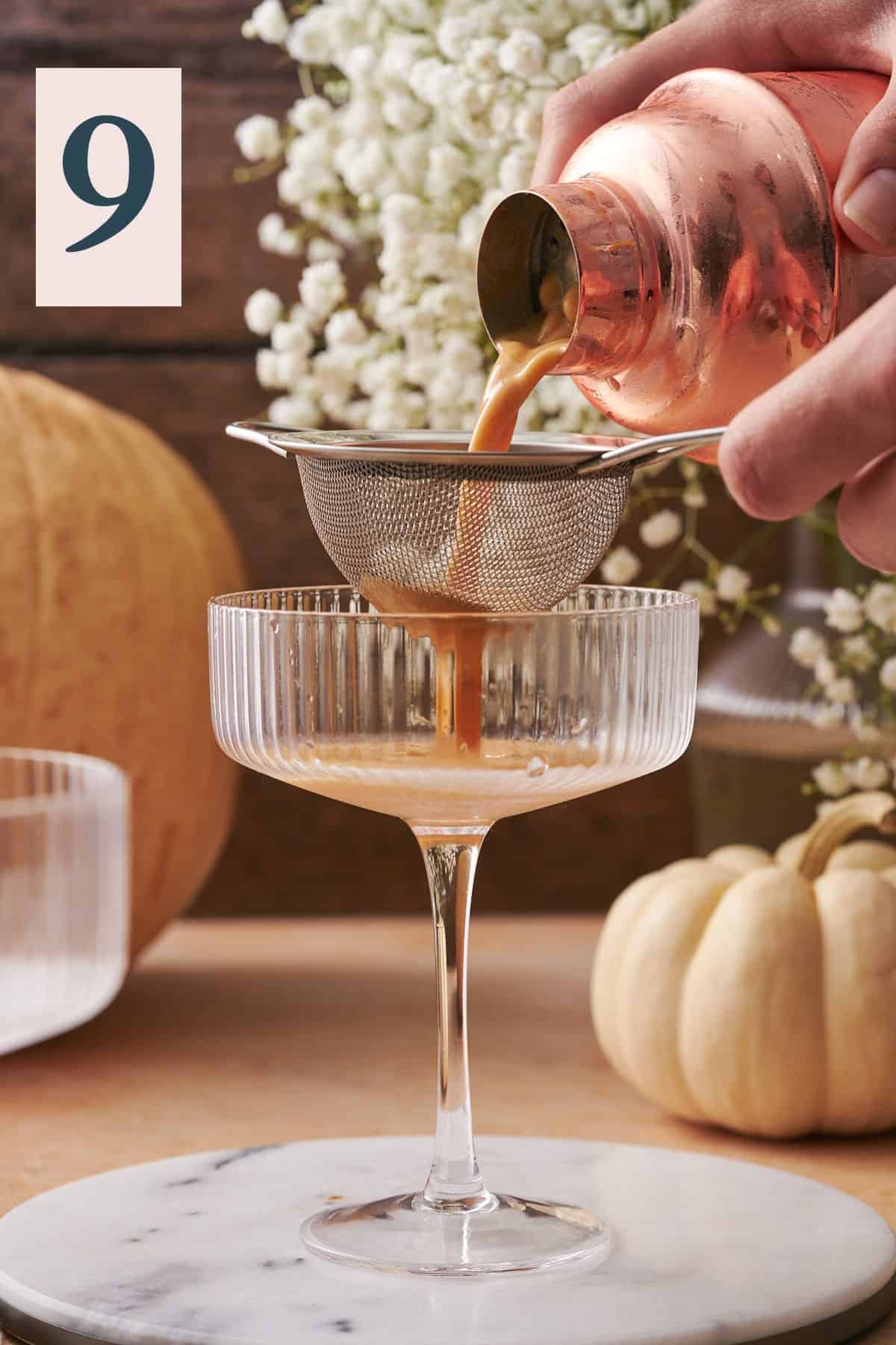 hand pouring pumpkin martini mixture into a martini glass through a metal sieve. 