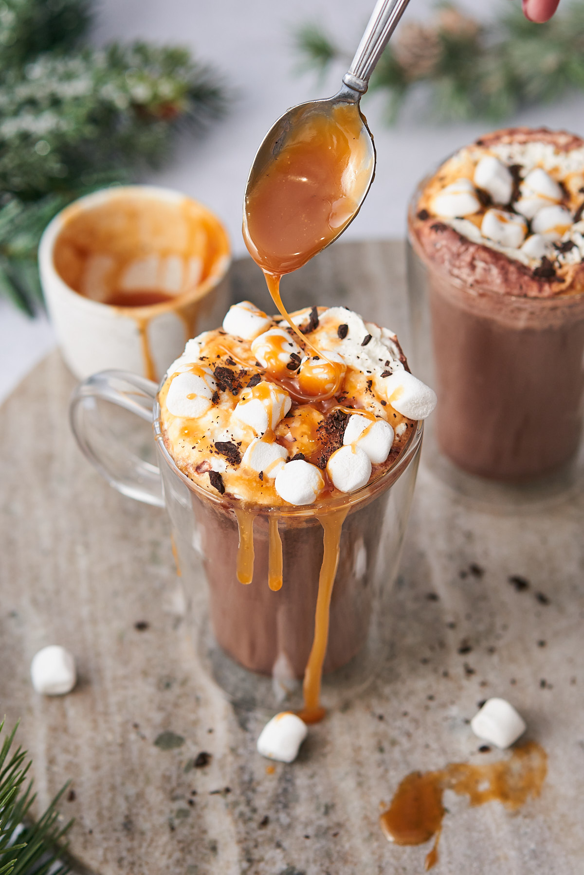 hot cocoa topped with marshmallows, whipped cream, chocolate shavings, and a spoonful of salted caramel being drizzled on top. 