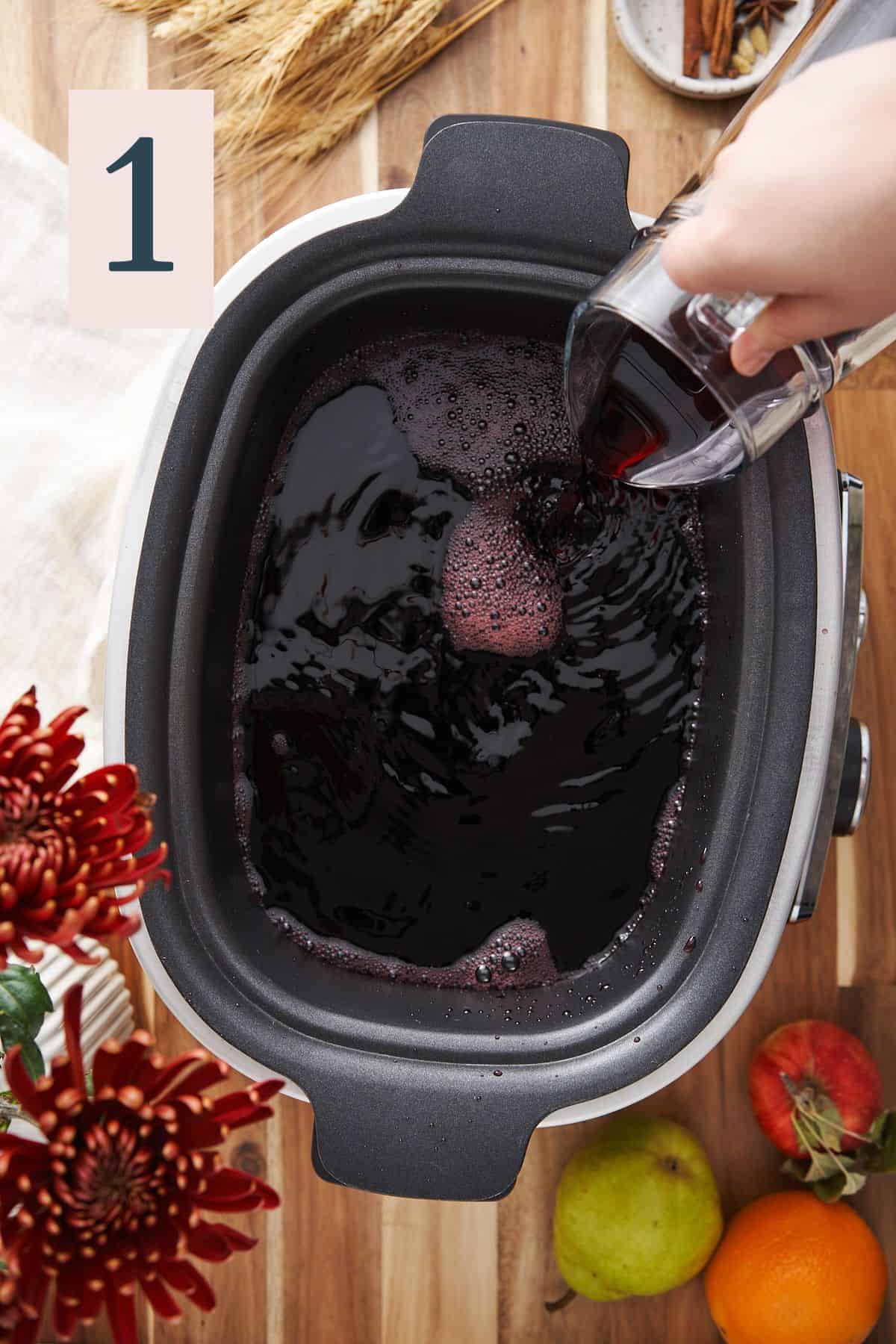 hand pouring red wine from glass pitcher into a large slow cooker, surrounded by red mums and fresh fruit.