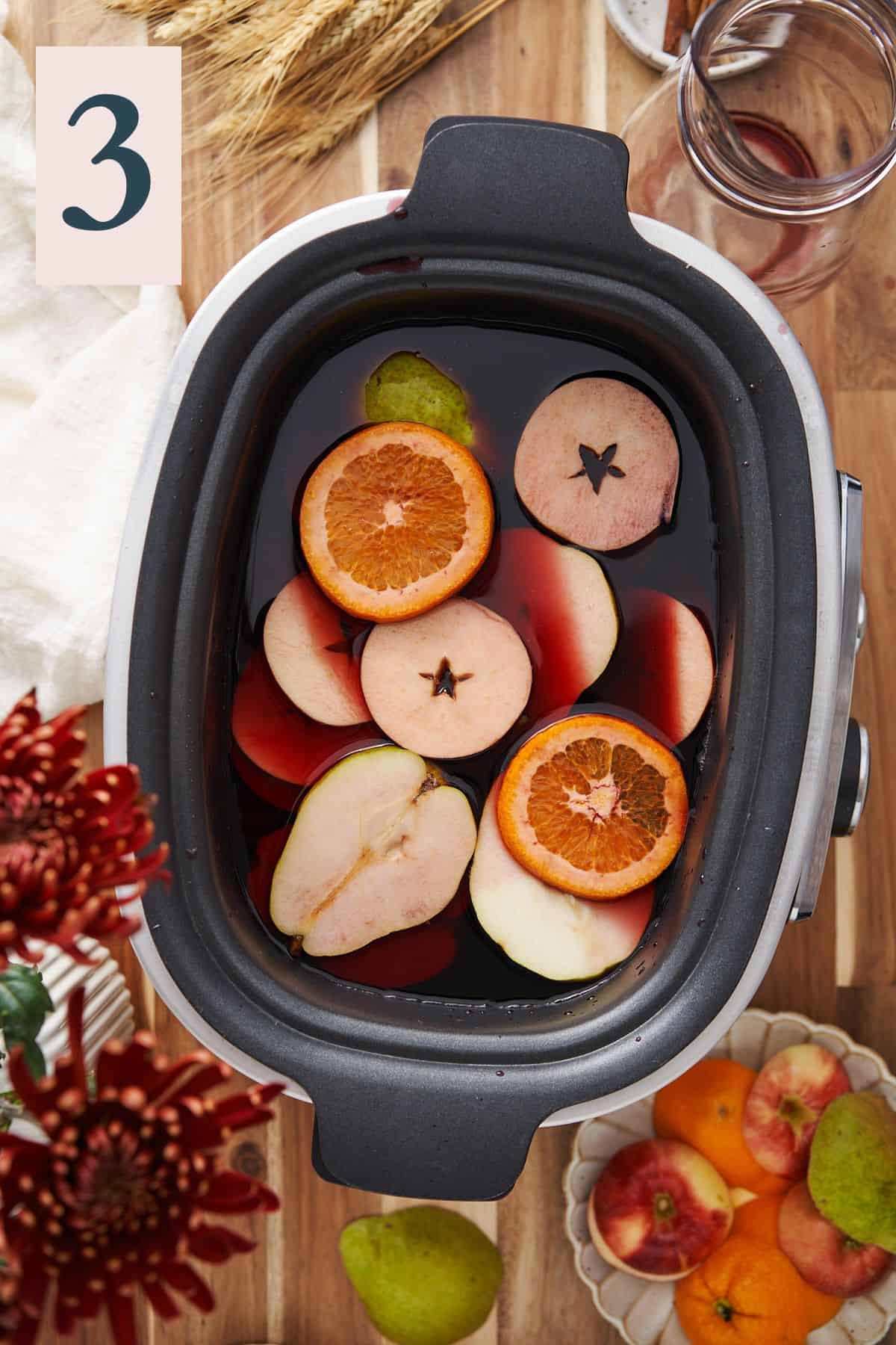 orange and apple rounds with pears in a slow cooker with red wine, surrounded by fruit on a wood table. 
