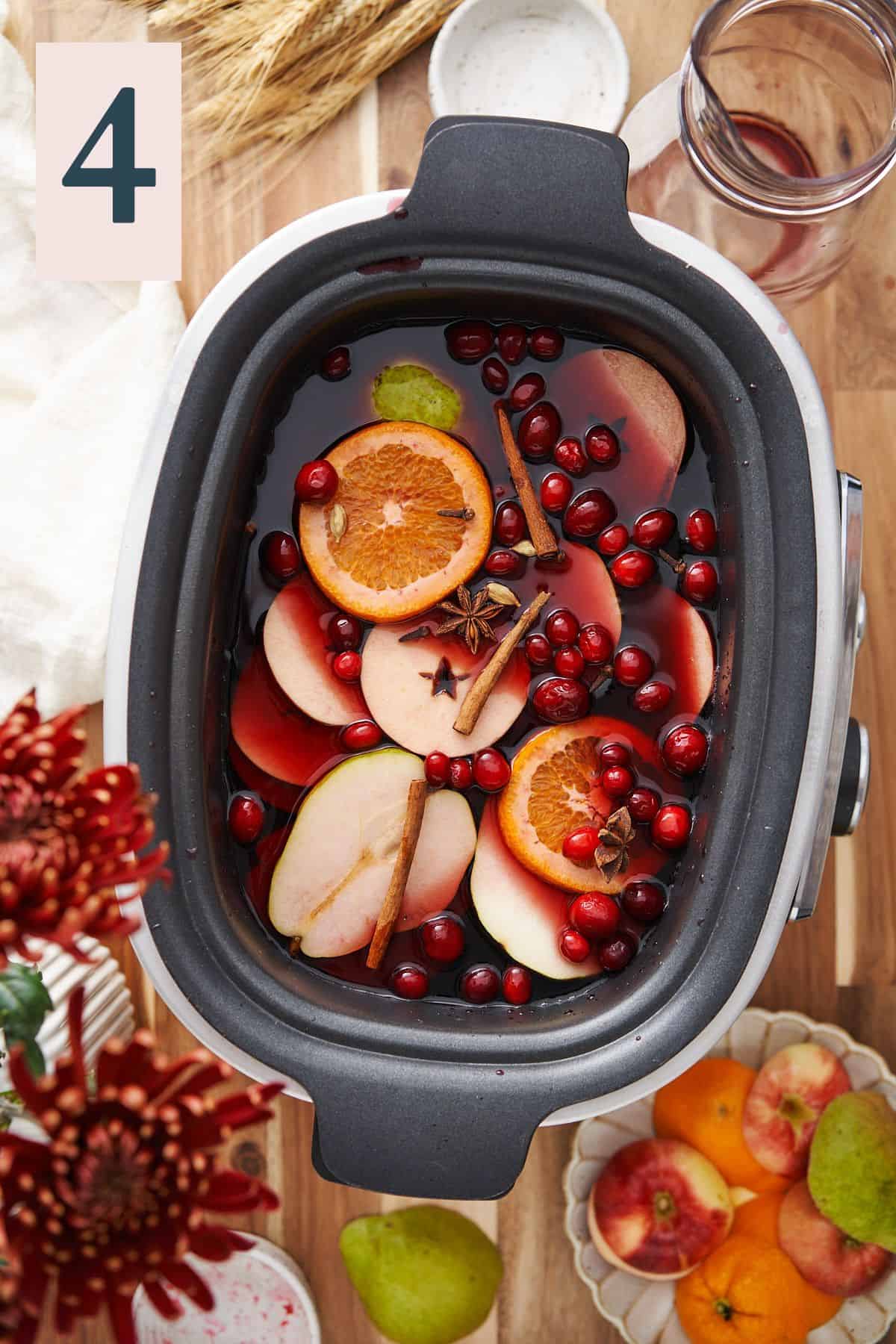 orange and apple rounds with pears, cranberries, cinnamon sticks, and other whole spices in a slow cooker with red wine, surrounded by fruit on a wood table. 