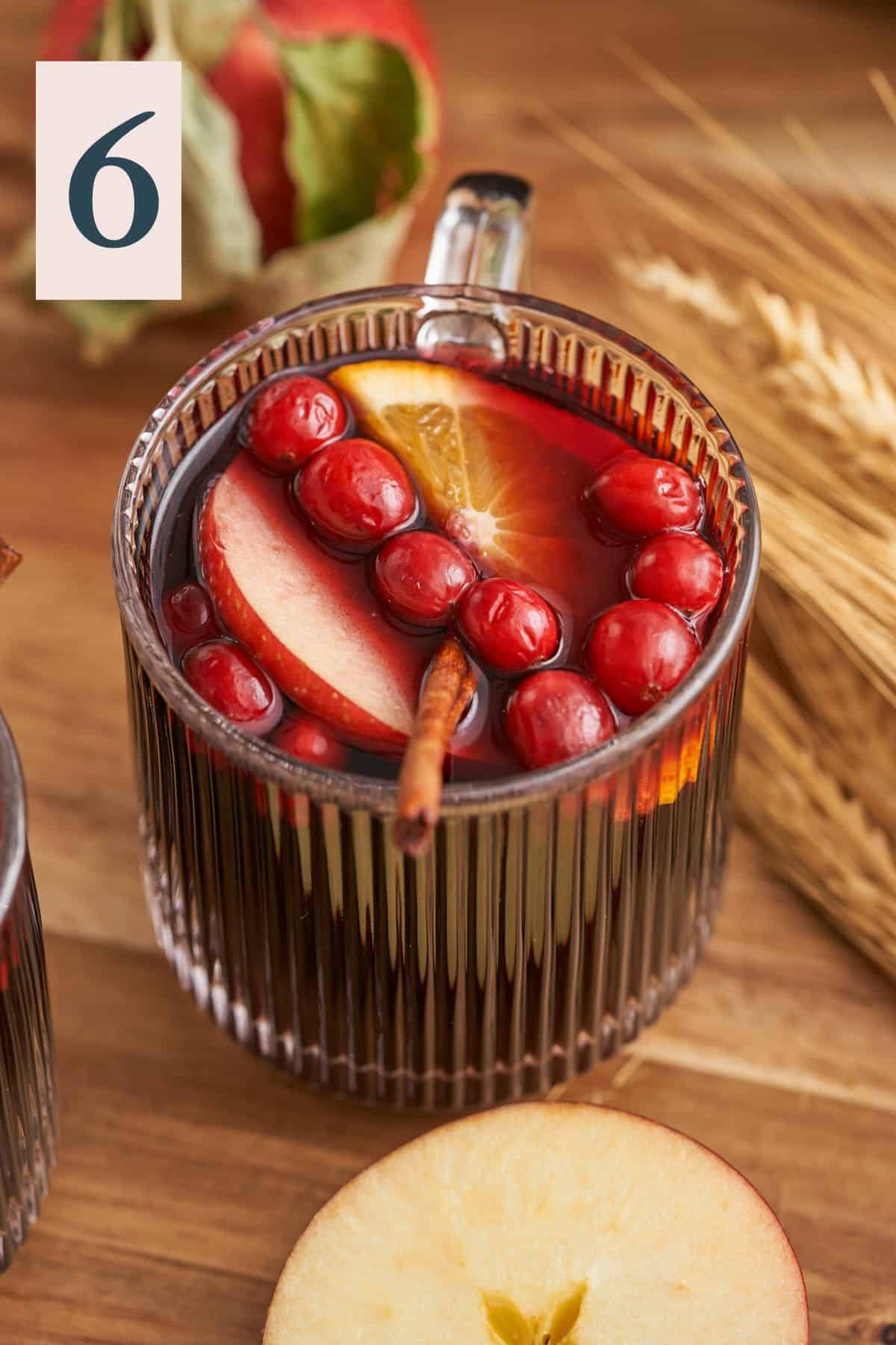 lovely cup full of mulled wine with fresh cranberries, apple slices, orange rounds and a cinnamon stick on a wood table with dried wheat stalks.