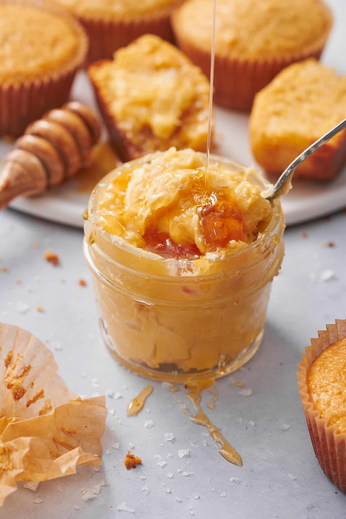 whipped honey butter in a small glass jar, with cornbread muffins in the background, and honey being drizzled on top. 