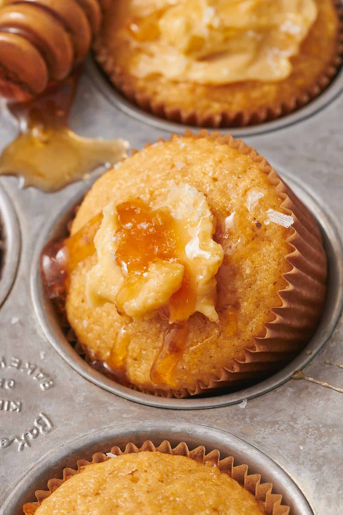 delicious honey cornbread muffins topped with whipped honey butter, sea salt, and honey on a silver baking tray with a honey dripper in the background.