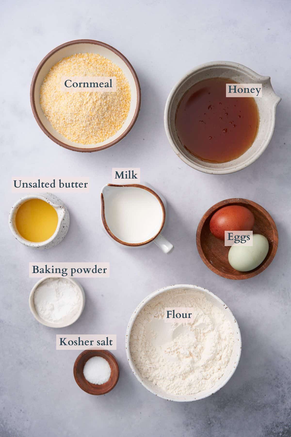 ingredients to make honey cornbread muffins laid out in wooden and ceramic bowls, labeled to denote each ingredient. 