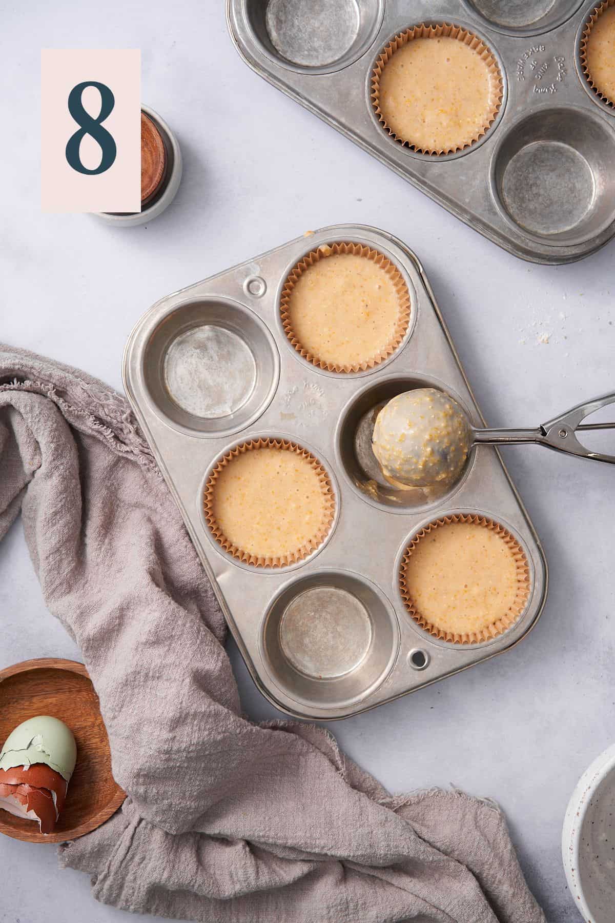 filling a muffin tin with cornbread muffin batter, and a large cookie scoop nearby the scene.