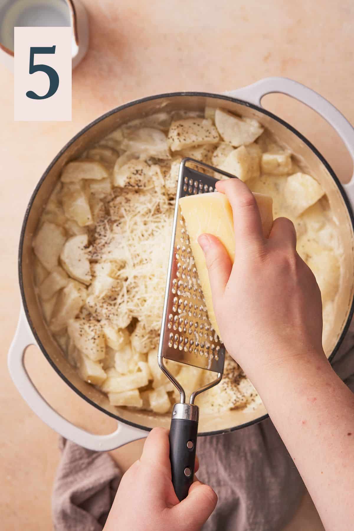 hand grating fresh parmesan into a dutch oven full of potatoes.