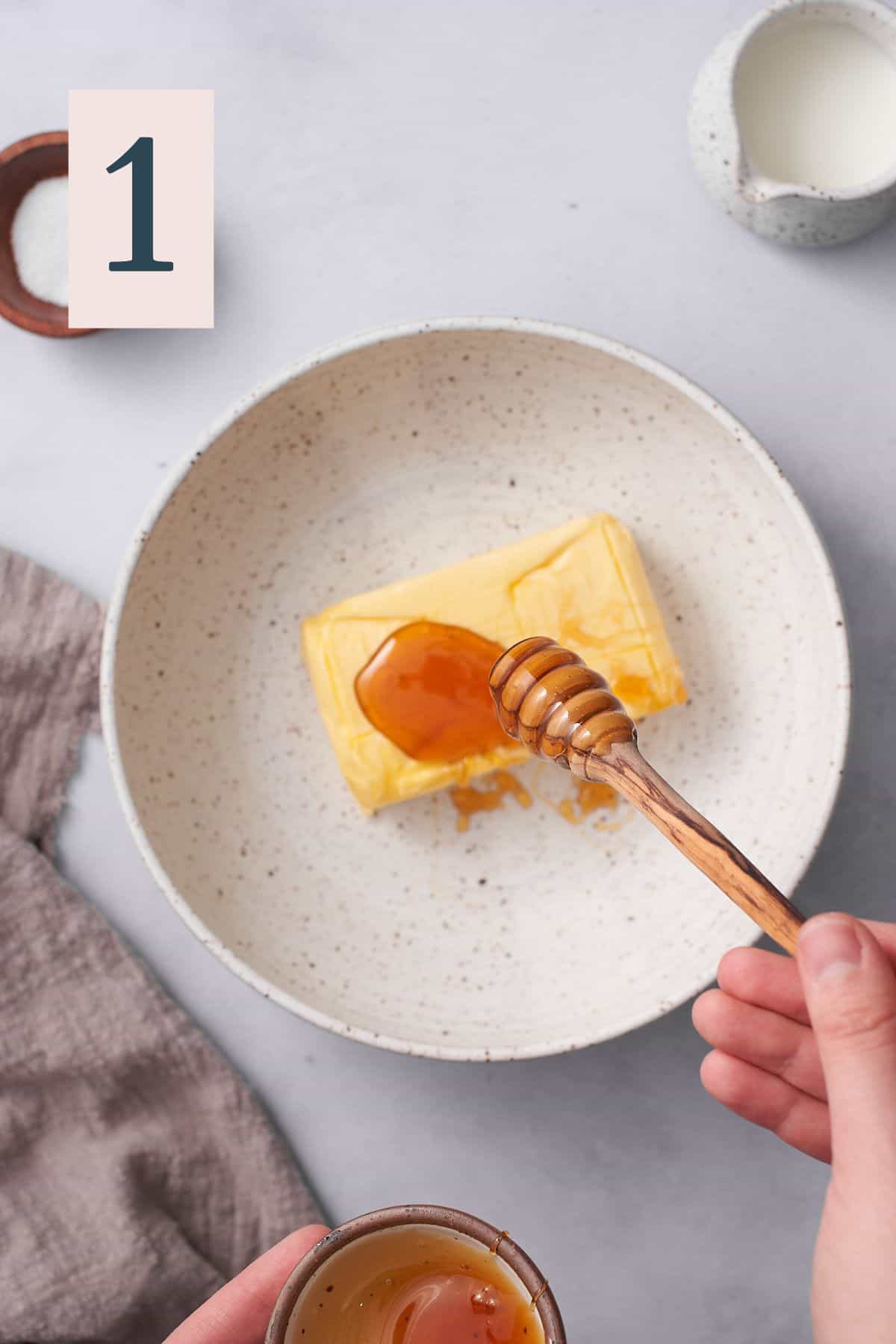 hand drizzling honey from a honey dripper onto block of butter in a mixing bowl. 