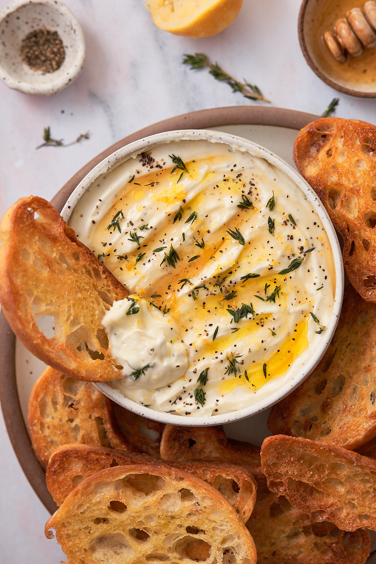 creamy whipped ricotta in a bowl, topped with honey, olive oil, fresh thyme, salt, and pepper, with crispy crostini surrounding it and one piece of bread dunked in the bowl. 