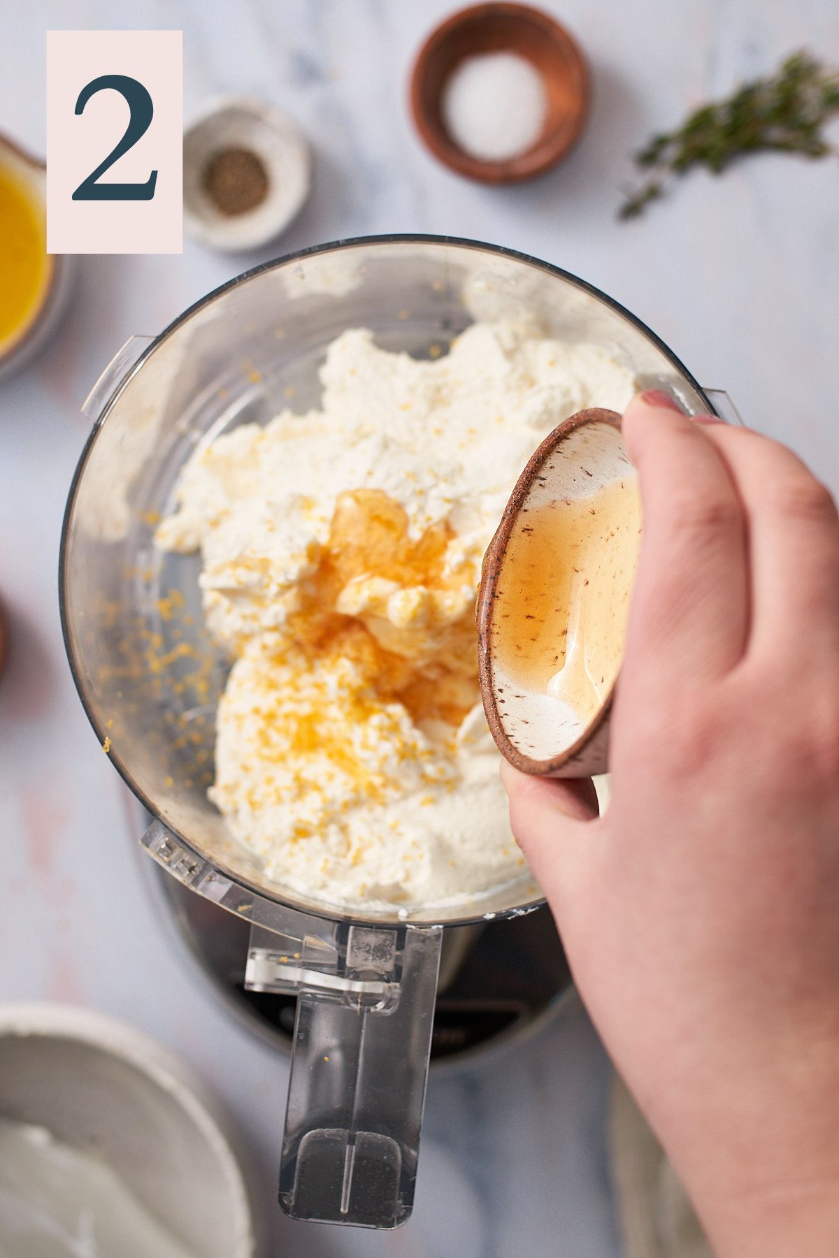 hand pouring honey into a food processor with ricotta and lemon zest. 