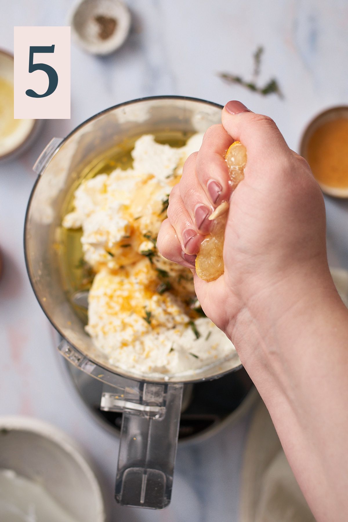 hand squeezing lemon juice into a food processor with ricotta, honey, olive oil, lemon zest, spices and fresh thyme.