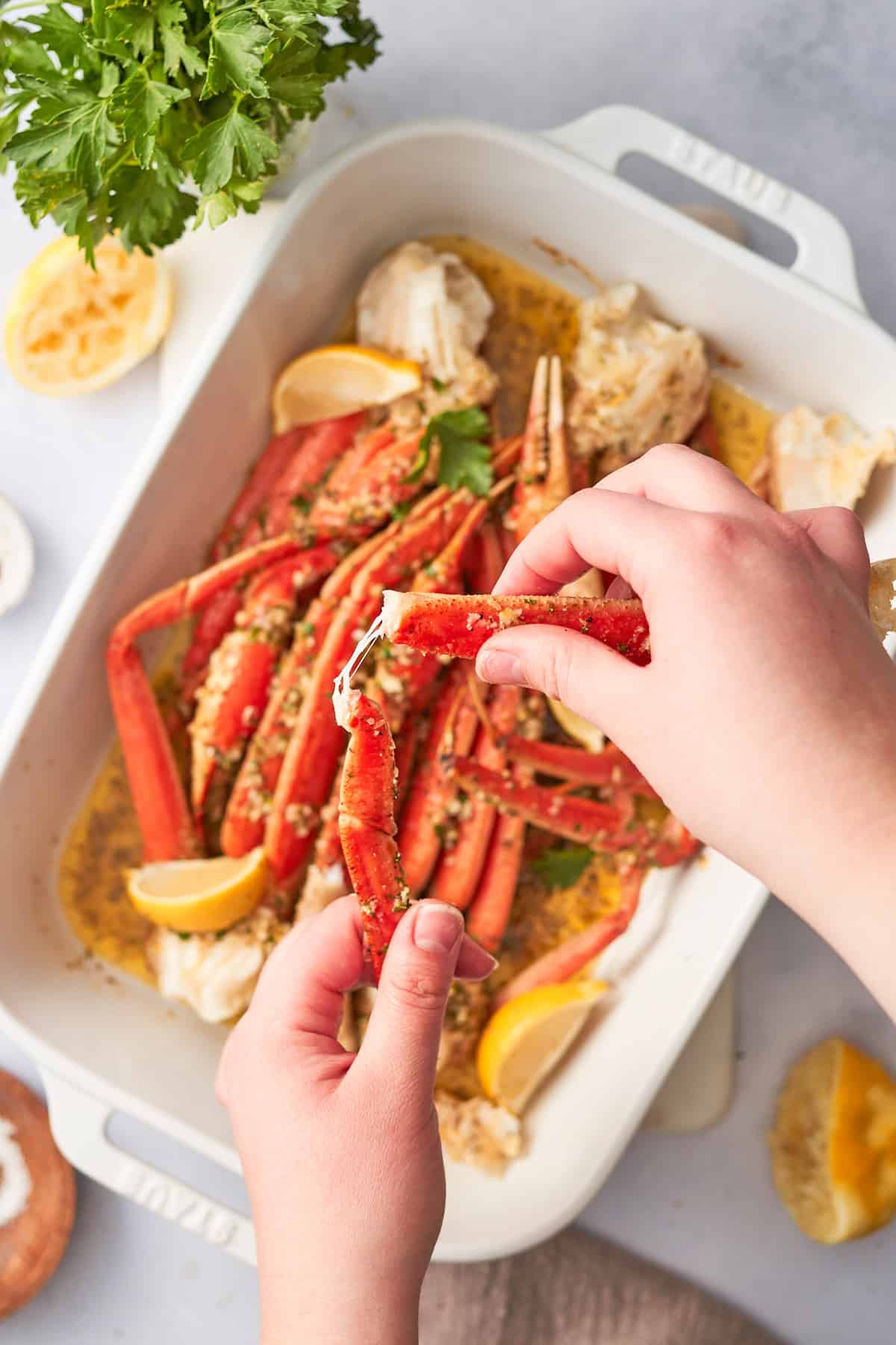Hands demonstrating how to crack a crab leg at the joint over a baking dish full of crab legs with buttery lemon garlic butter.