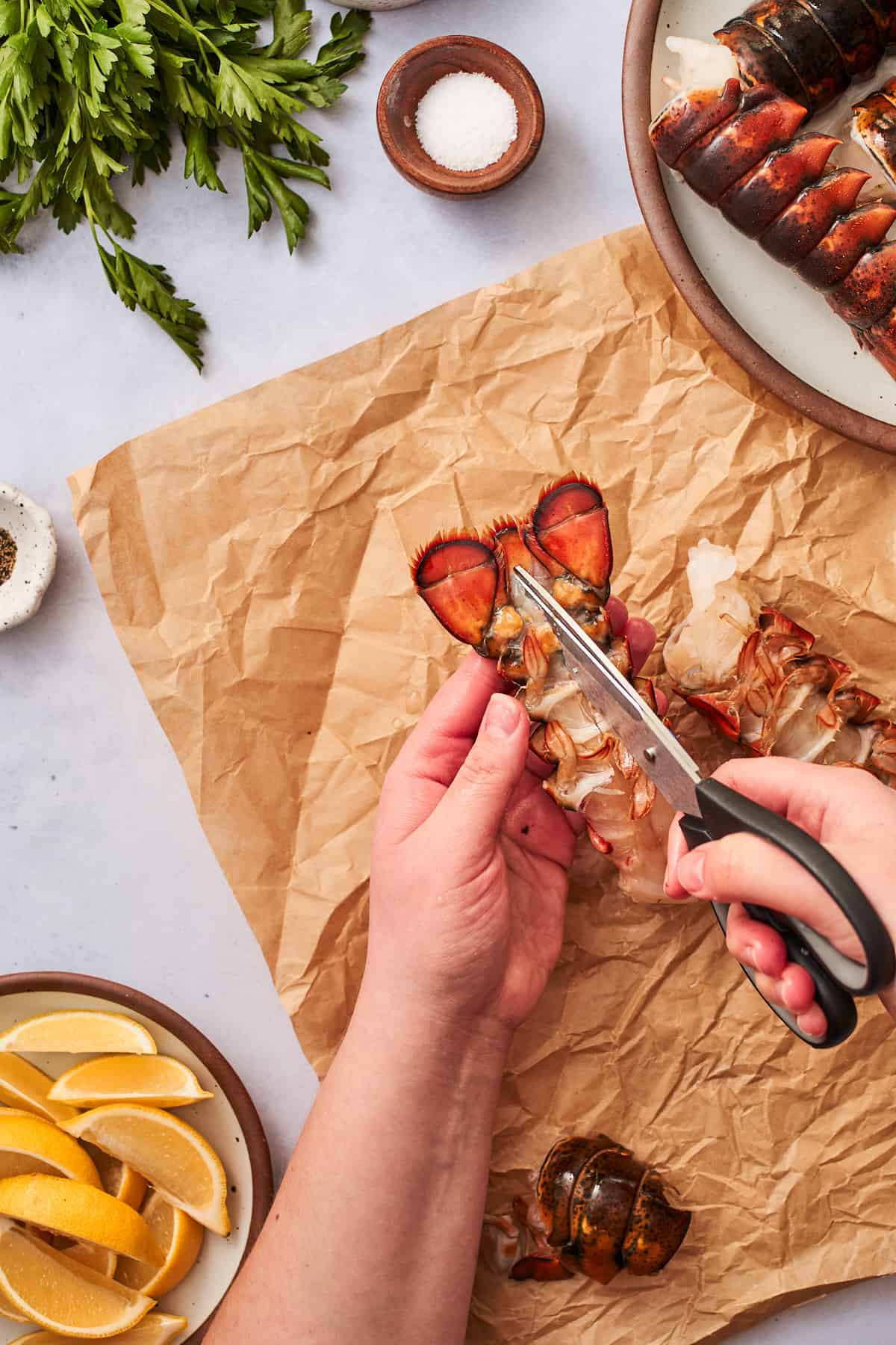 Hand cutting lobster tail from the underside with a pair of kitchen shears, with parchment paper underneath.