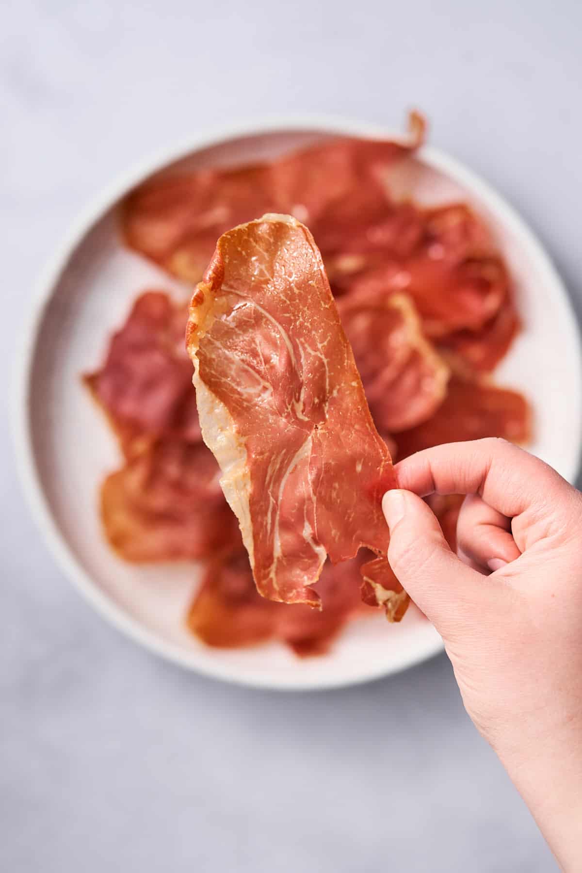 hand holding a slice of prosciutto to the camera, with prosciutto on a plate below it. 
