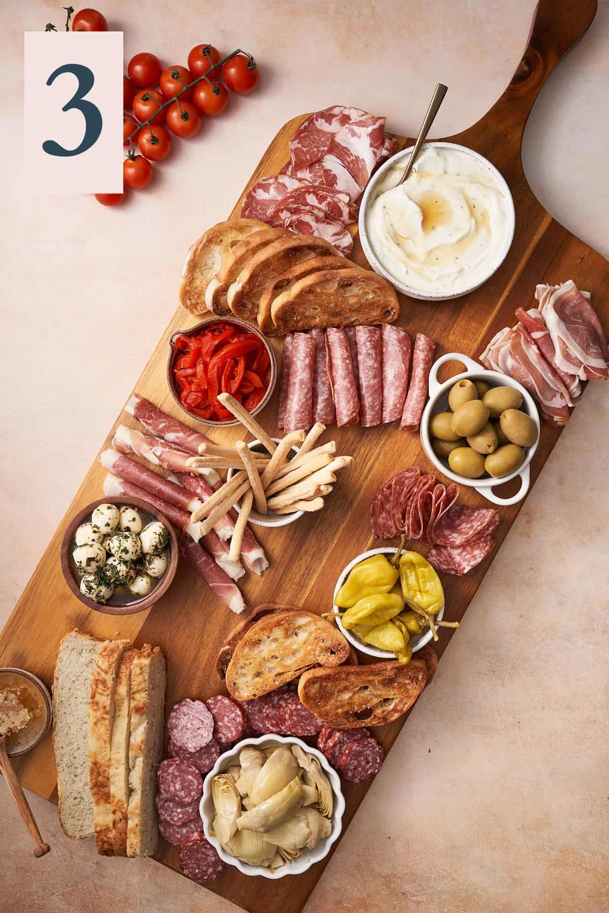 breads, meats, cheeses, olives, and peppers on a large wooden board. 