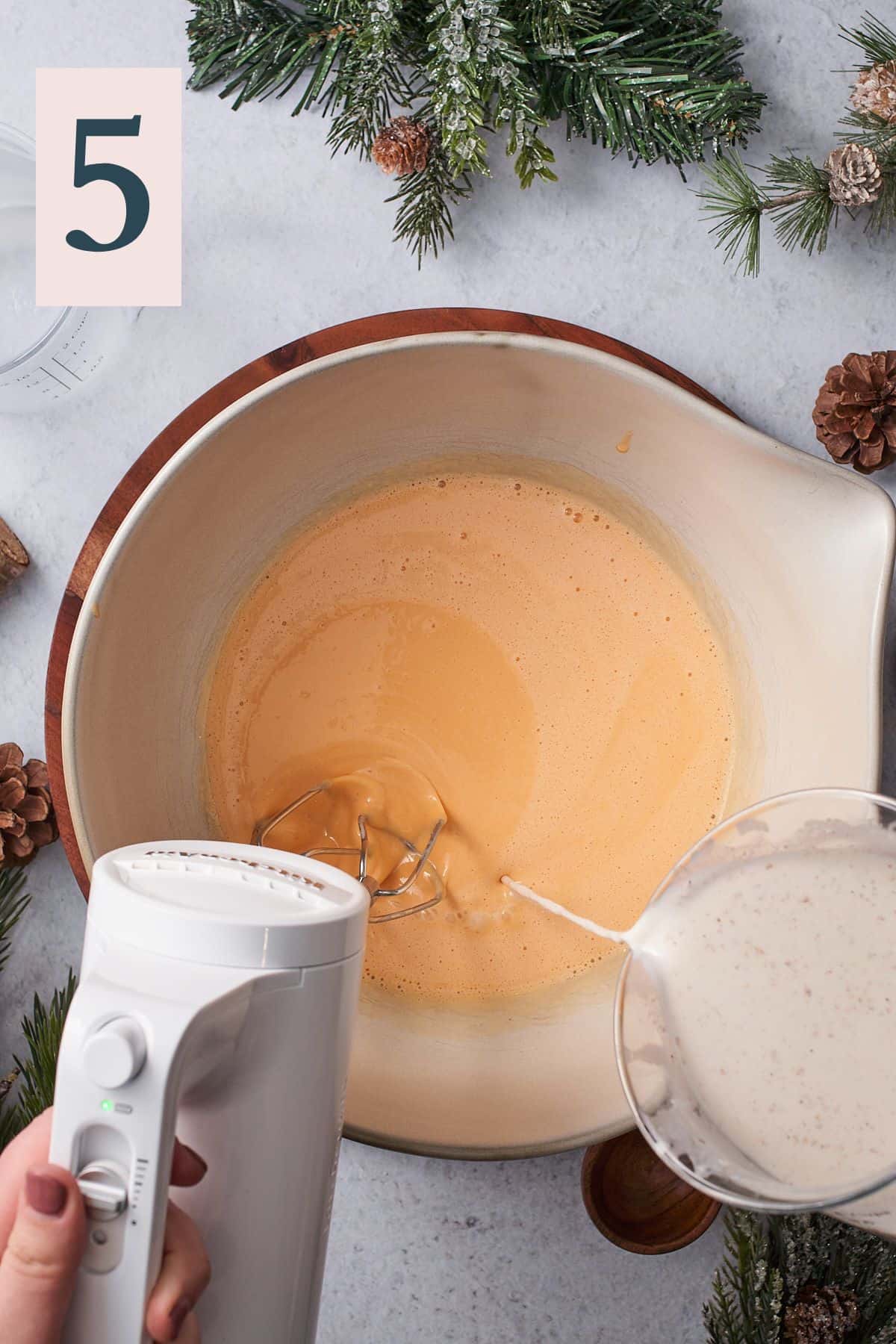 hand pouring spiced milk mixture into a bowl of creamy egg yolks, with a hand mixer to mix everything together. 