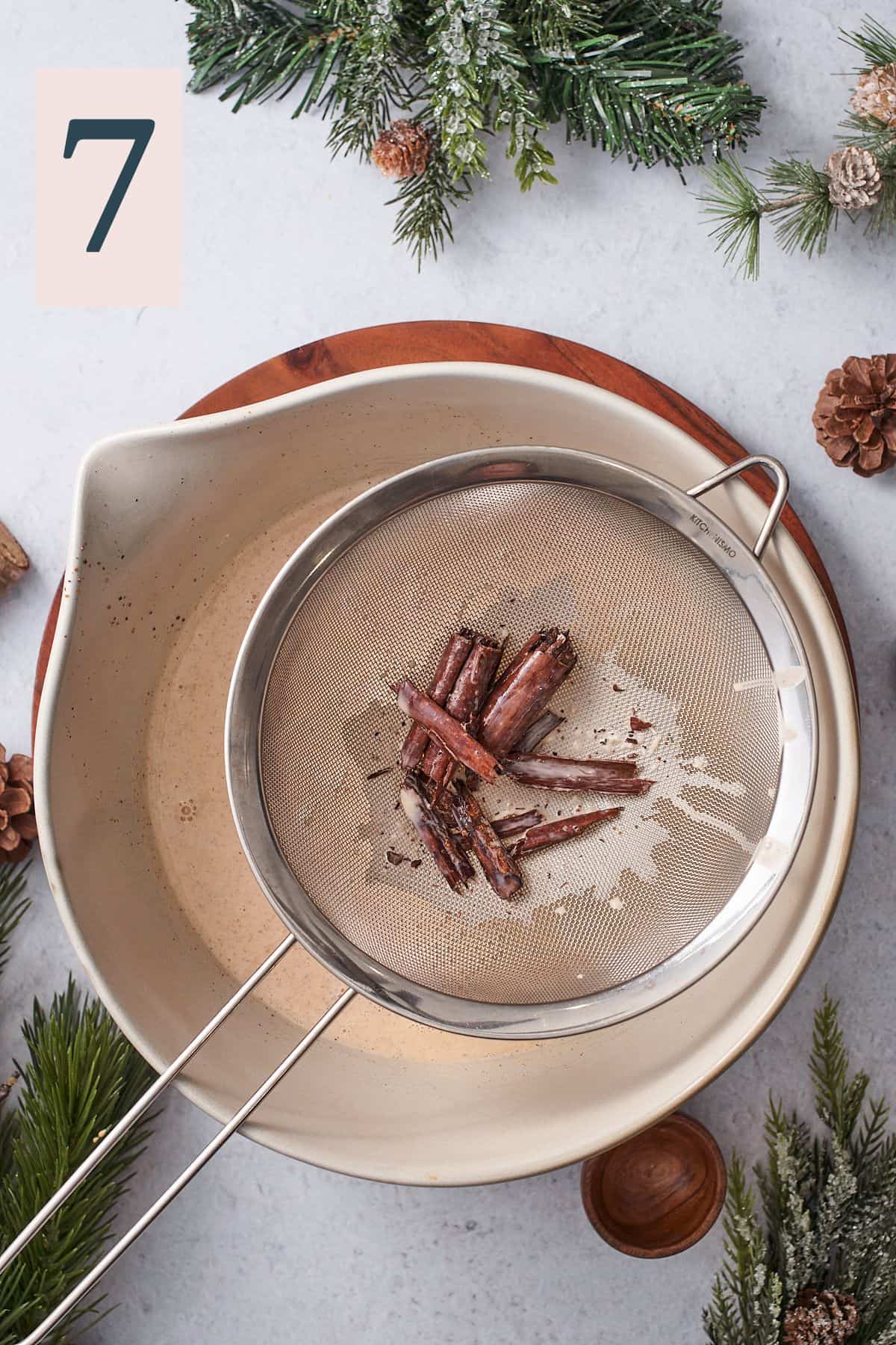 straining cinnamon sticks out of an eggnog mixture in a metal sieve over a large bowl. 