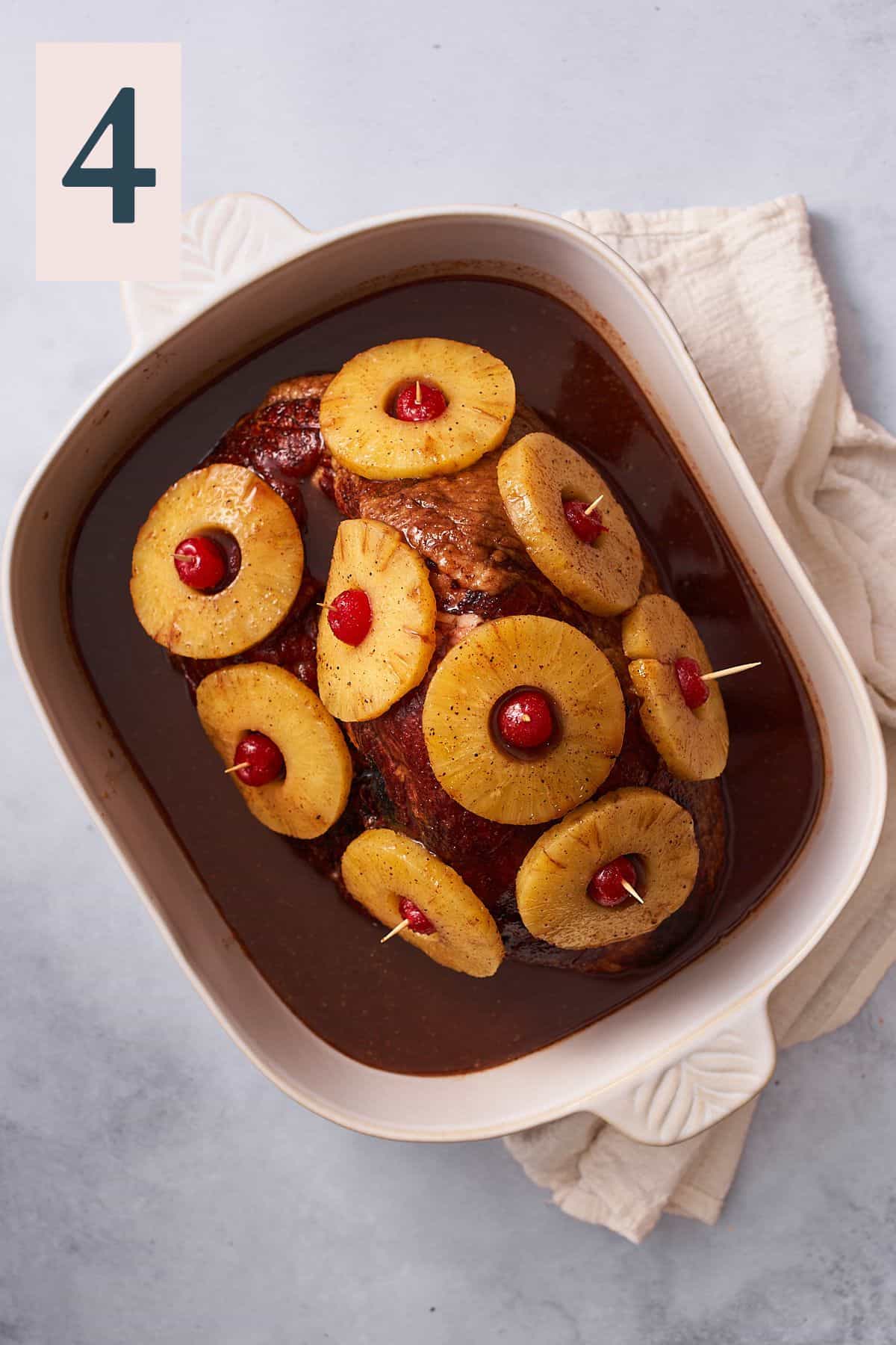ham covered in pineapple rings with maraschino cherries in a roasting pan with liquid at the bottom.