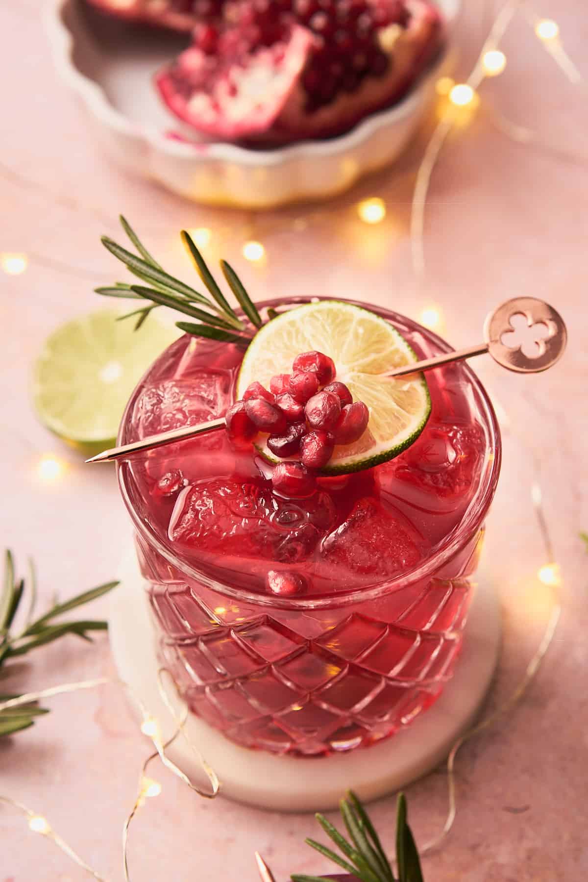 a stunning bright red pomegranate gin cocktail with rosemary, lime, and pomegranate arils, surrounded by tiny twinkling fairy lights. 